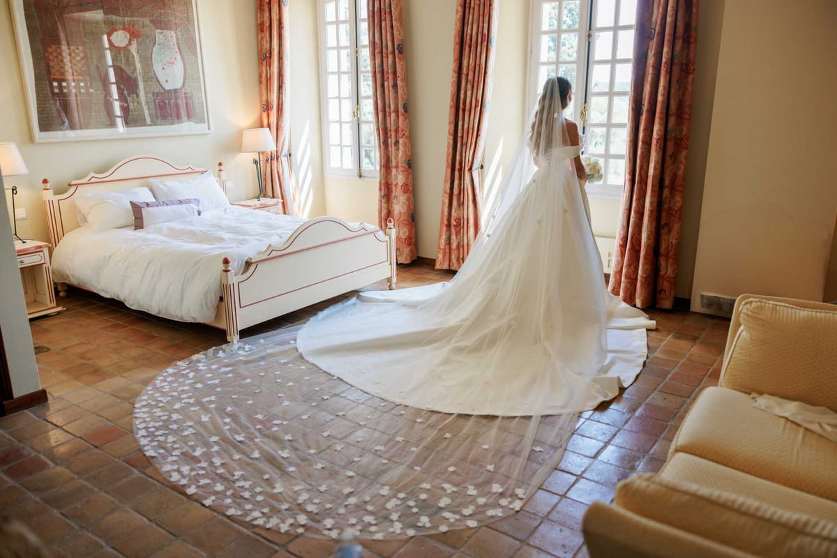A bride stands facing an open window in a furnished bedroom, photographed from behind in a wide shot that showcases the full length of her ivory off-the-shoulder ball gown. The cathedral-length veil is heavily embellished with small three-dimensional floral appliqués scattered across its entire surface, fanning out dramatically across the terracotta tile floor. The room features a white painted wooden bed frame with red detailing, white bedding with lavender accent pillows, floral-patterned coral and salmon curtains, warm lamp lighting, and a large framed artwork on the wall. The bride holds a small white bouquet and the overall interior style is classic French country.