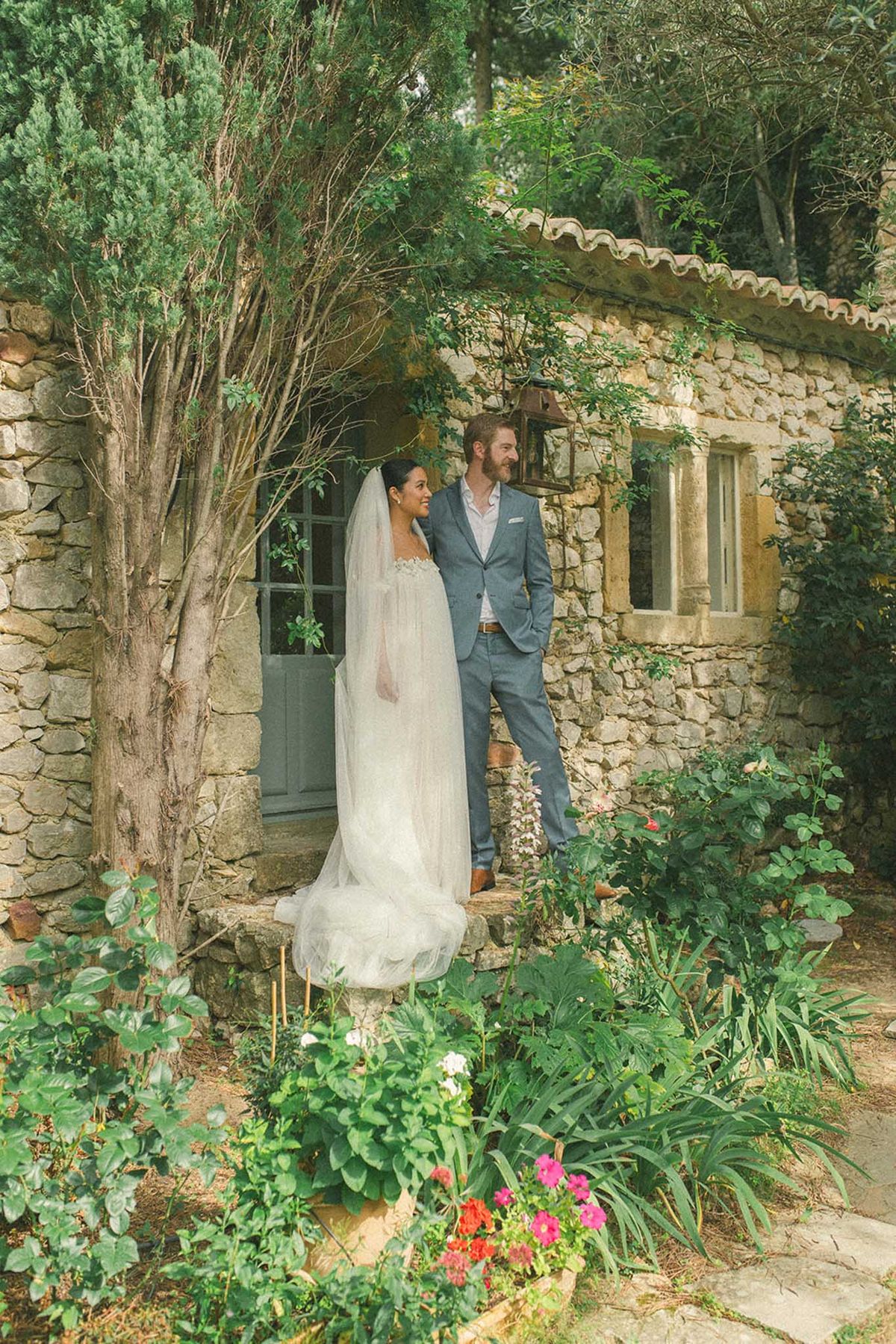 Bride and groom posing outside stone cottage with ivy-covered walls and flowering garden in foreground