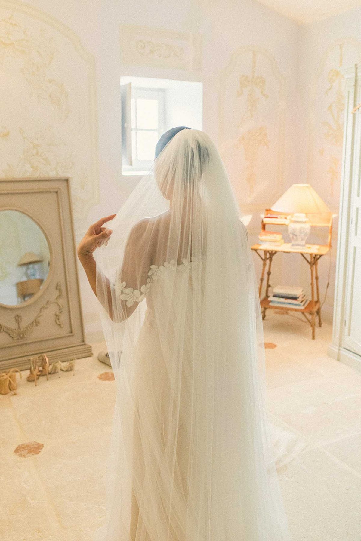 Bride standing with flowing veil and blue headpiece in elegant bedroom with damask wallpaper