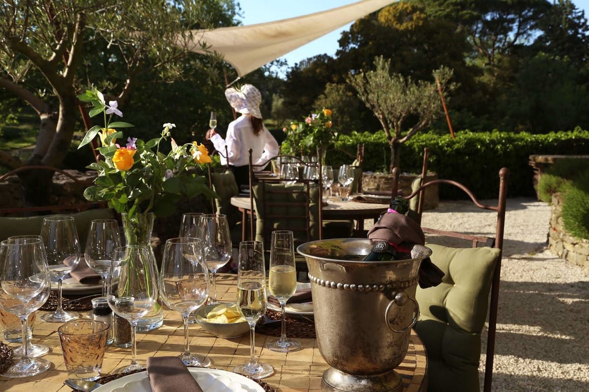 An outdoor wedding reception or cocktail hour setup in a garden setting, featuring round tables arranged on a gravel terrace shaded by a cream canvas sail shade. The foreground table is set with multiple wine glasses and champagne flutes, taupe linen napkins, amber-tinted glassware, dark wicker placemats, and a loose floral arrangement of yellow and orange roses with small purple flowers in a clear glass vase. A silver beaded champagne bucket holds a chilling bottle wrapped in a brown cloth. Chairs are fitted with olive green cushions and have dark wrought-iron frames. One woman, seen from behind and wearing a white top and a floral bucket hat, stands holding a champagne flute near the tables in the middle ground. The decor palette is warm and natural — earthy tones, aged metals, and garden greenery — consistent with a rustic Provençal or countryside style. Wide shot with a shallow depth of field, keeping the foreground table sharp and the background softly blurred.