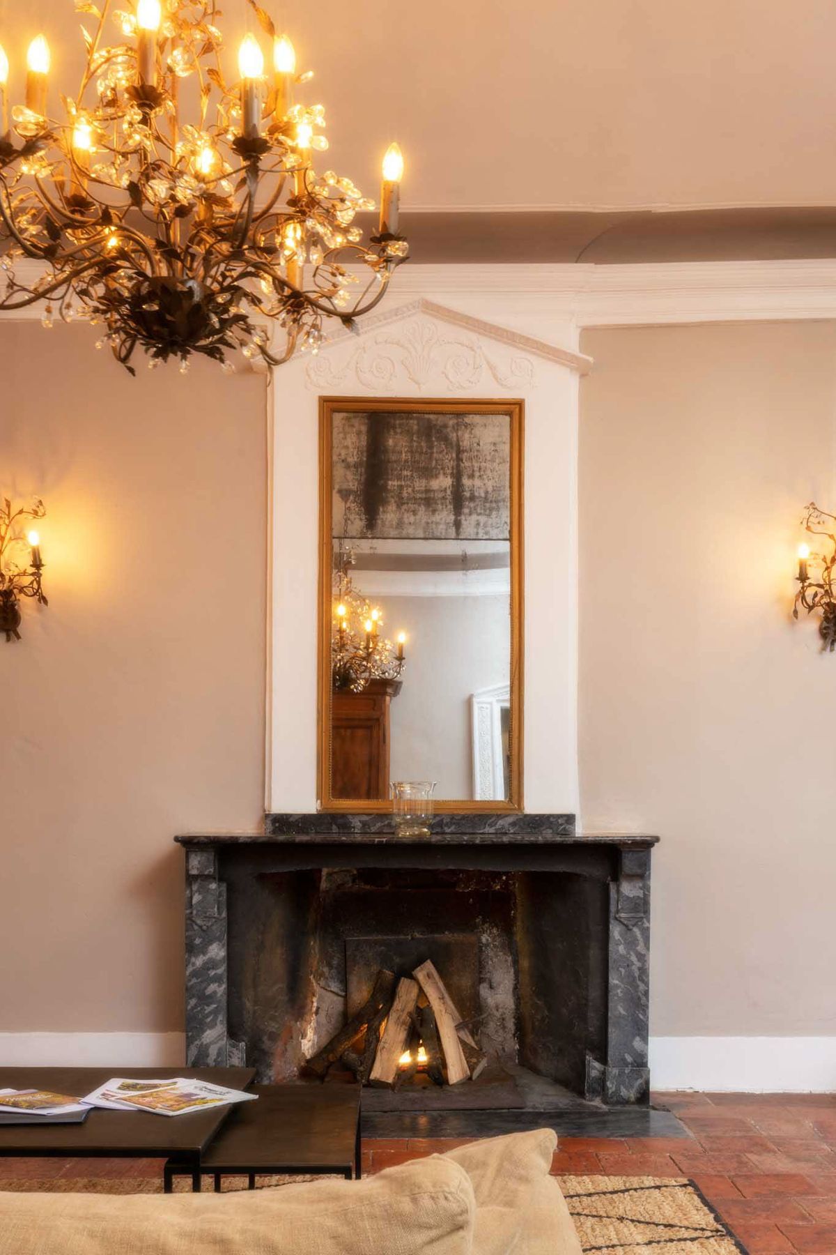 Interior shot of a château reception room featuring a dark grey marble fireplace with logs set inside, topped by a tall gold-framed antique mirror set within an ornate white plasterwork alcove with carved decorative detailing. The room is lit by a wrought-iron branch-style chandelier with crystal accents and candle-style bulbs in the upper left, and matching wall sconces on either side of the fireplace. The walls are painted in a warm off-white or pale blush tone, and the floor is terracotta tile partially covered by a natural woven rug. A low black coffee table with magazines and a cream upholstered seat are visible in the foreground. No people are present. Potential venue feature image.
