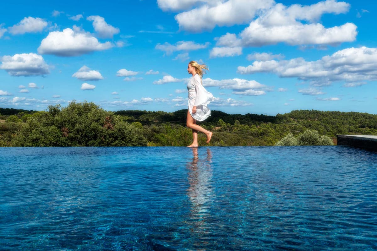 Barefoot woman in white flowing dress walking at edge of infinity pool overlooking green woodland