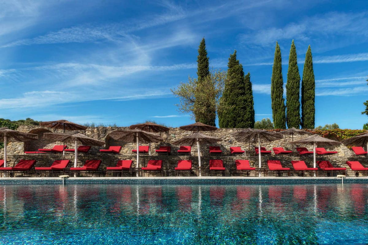 Swimming pool with red-cushioned loungers under straw parasols backed by stone wall and cypress trees