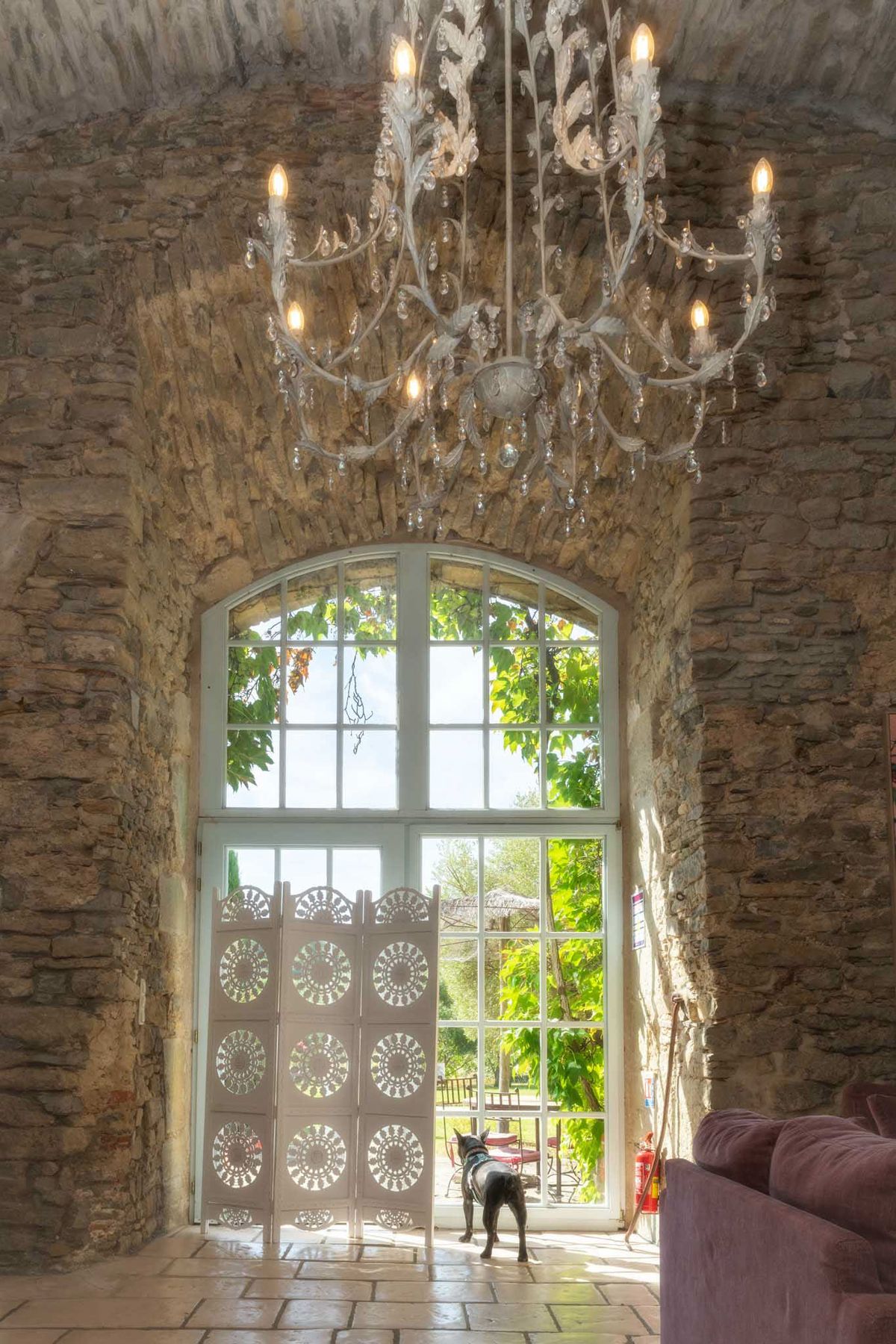 Interior architectural shot of a rustic French venue space featuring exposed stone walls with a vaulted arch framing a tall white-framed arched window that looks out onto a garden. A white ornate chandelier with crystal droplets and candle-style bulbs hangs prominently from the ceiling. In the foreground, a white decorative folding screen with circular cut-out patterns partially divides the space, and a small black dog stands near the base of the large window. A dusty mauve-pink sofa is partially visible on the right edge of the frame, and terracotta tile flooring is visible underfoot. Wide interior shot with natural backlight coming through the window. Potential venue feature image.