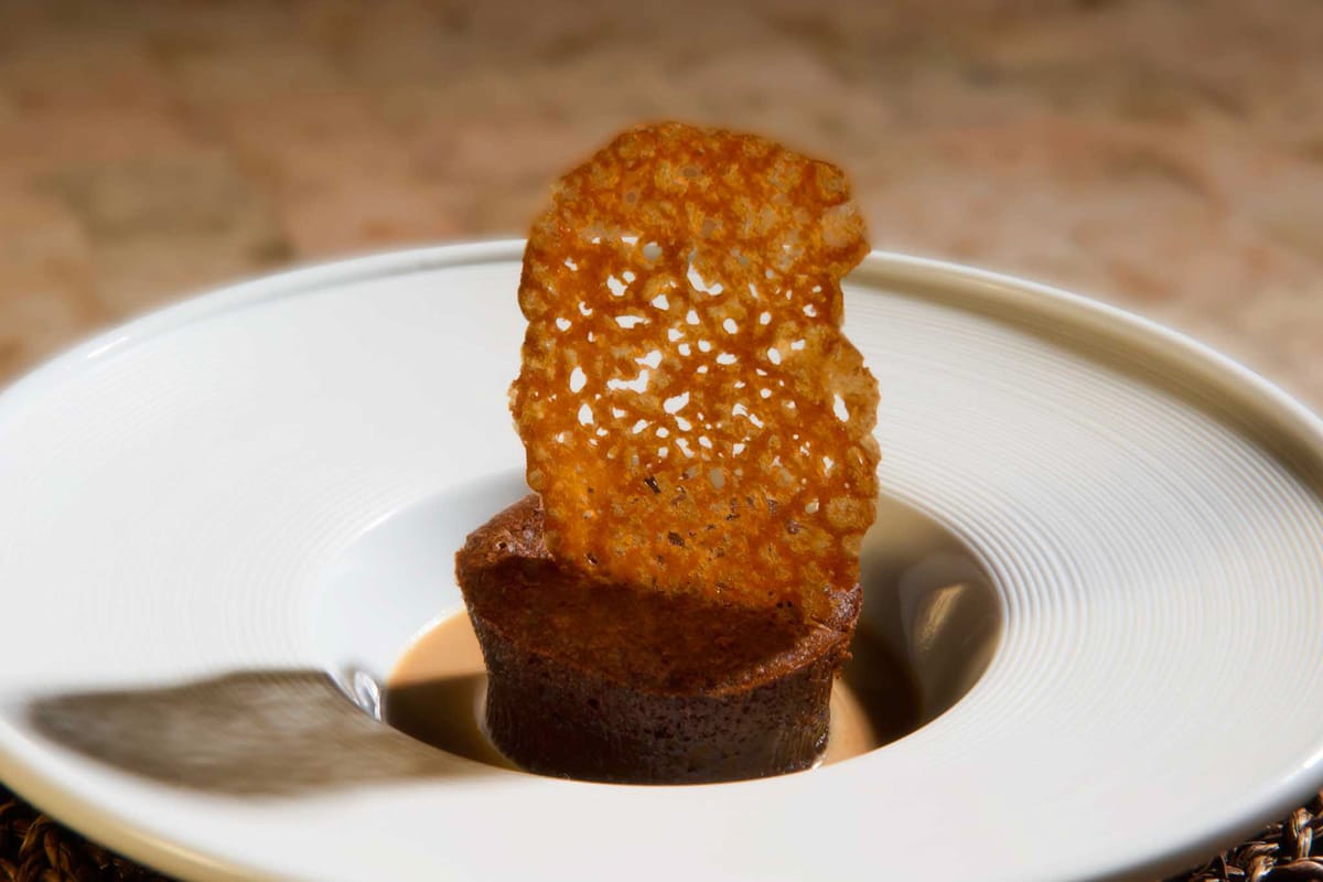 This image shows a close-up detail shot of a plated dessert course, likely from a wedding reception dinner service. The dish features a small dark chocolate tart or fondant base topped with a lace-patterned caramel or tuile wafer, served in a white ridged bowl with a spoon visible on the side. The presentation style suggests a formal, upscale catering aesthetic with attention to plated dessert details. This is a food detail photograph rather than a scene depicting people, a couple, or a wedding setting.