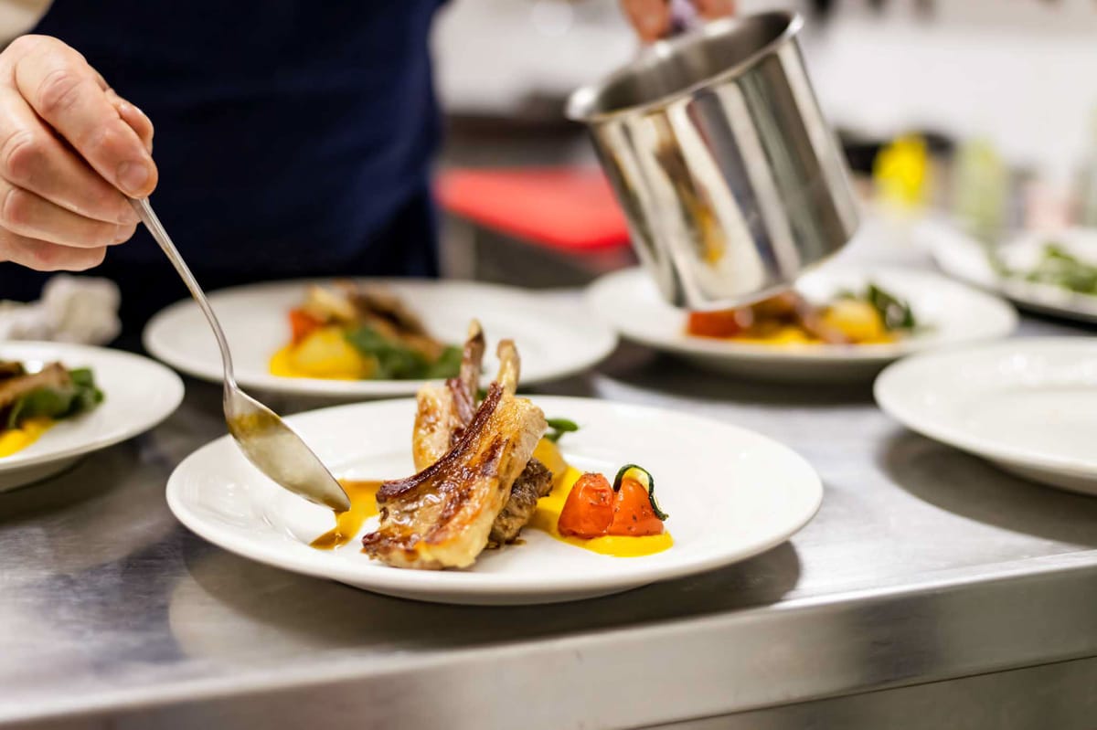 Chef plating roasted meat with yellow sauce and vegetables on stainless steel pass