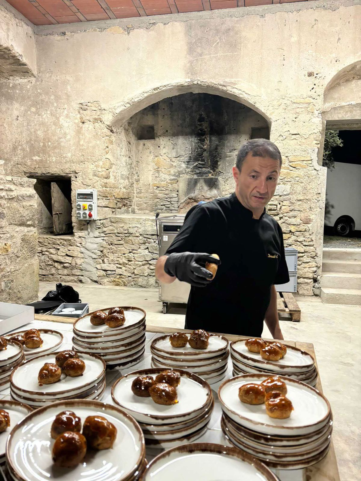 A caterer or chef in a black uniform and black gloves is plating wedding desserts in what appears to be an outdoor or semi-open kitchen area within a rustic stone-walled venue. He is arranging glazed profiteroles or choux pastry balls onto multiple stacked white plates with brown-rimmed edges, with numerous plates already prepared and lined up across a wooden work surface. The setting features exposed stone walls with a large arched alcove showing fire-blackened marks, suggesting a historic former bread or wood-fired oven, along with visible commercial kitchen equipment in the background. The image is a medium close-up shot capturing the food preparation and service workflow during what appears to be a wedding reception dinner service.