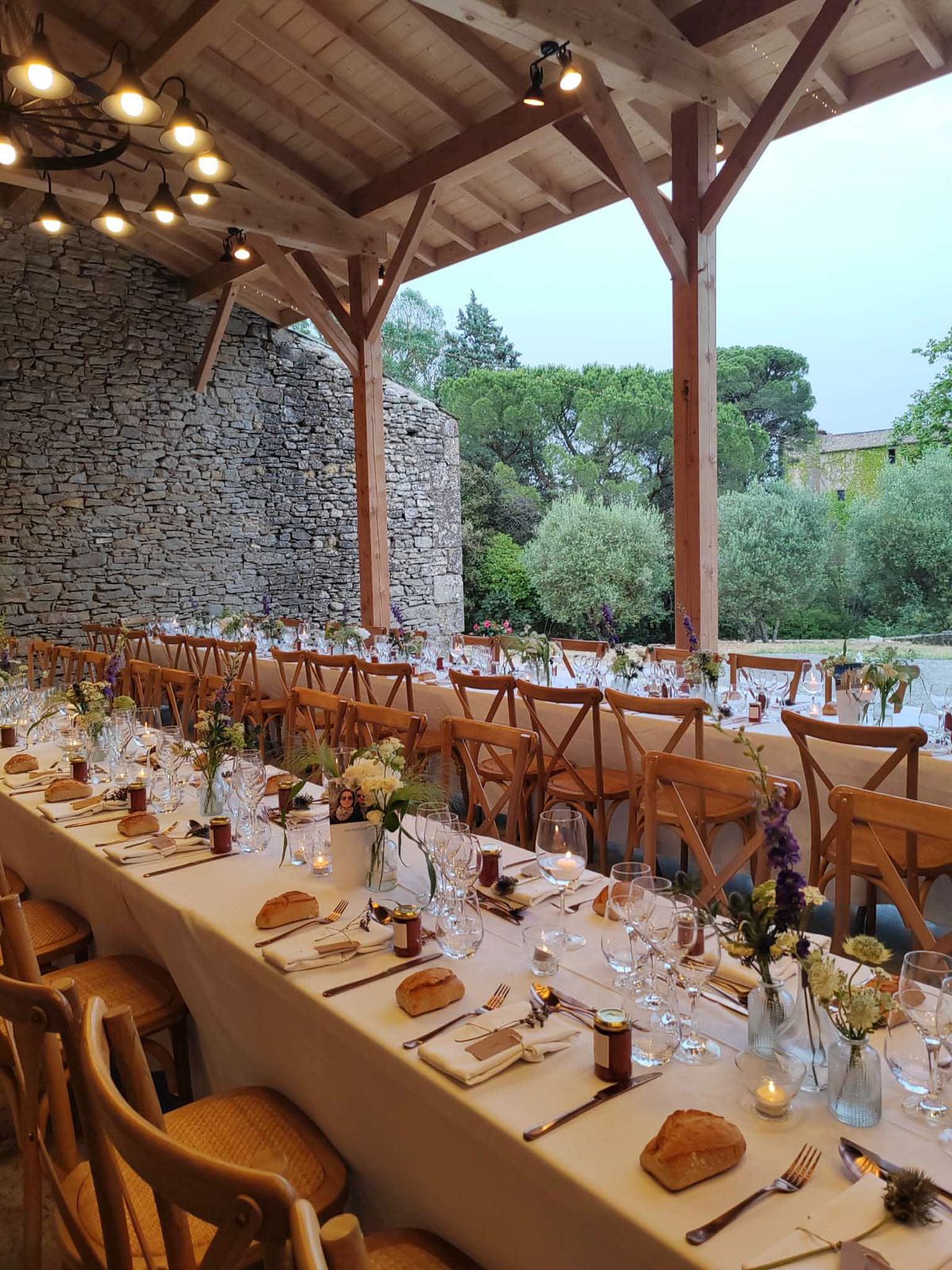 Pergola reception with Edison lights wildflower bud vases amber votives cross-back chairs and stone wall at dusk