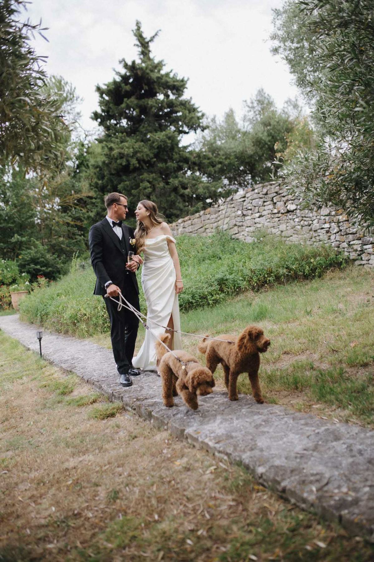 Bride and groom walking two dogs along stone garden path at a French chateau, groom holding champagne flute