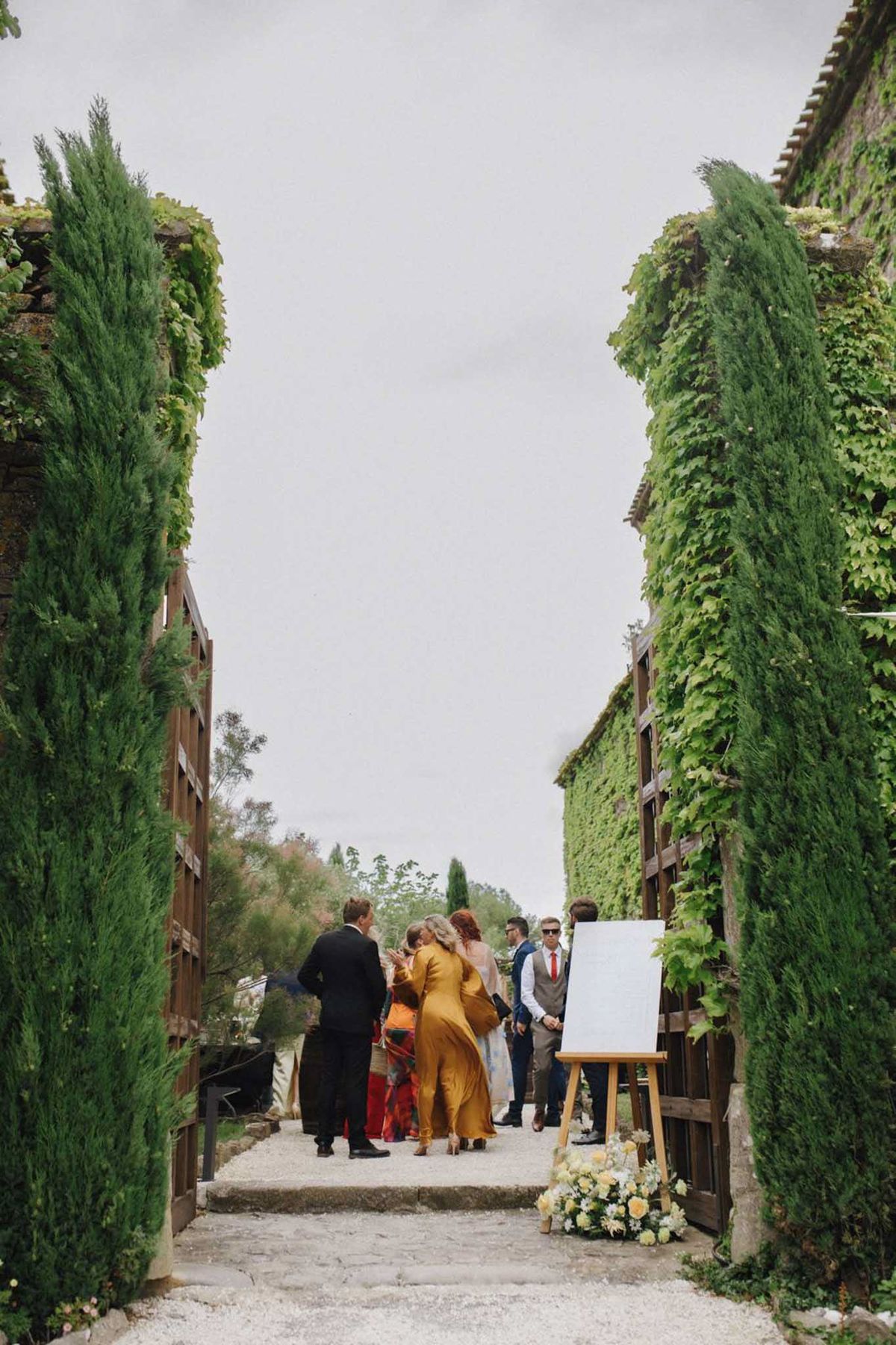 Six guests arriving along gravel pathway flanked by cypress trees welcome sign with yellow florals at venue entrance