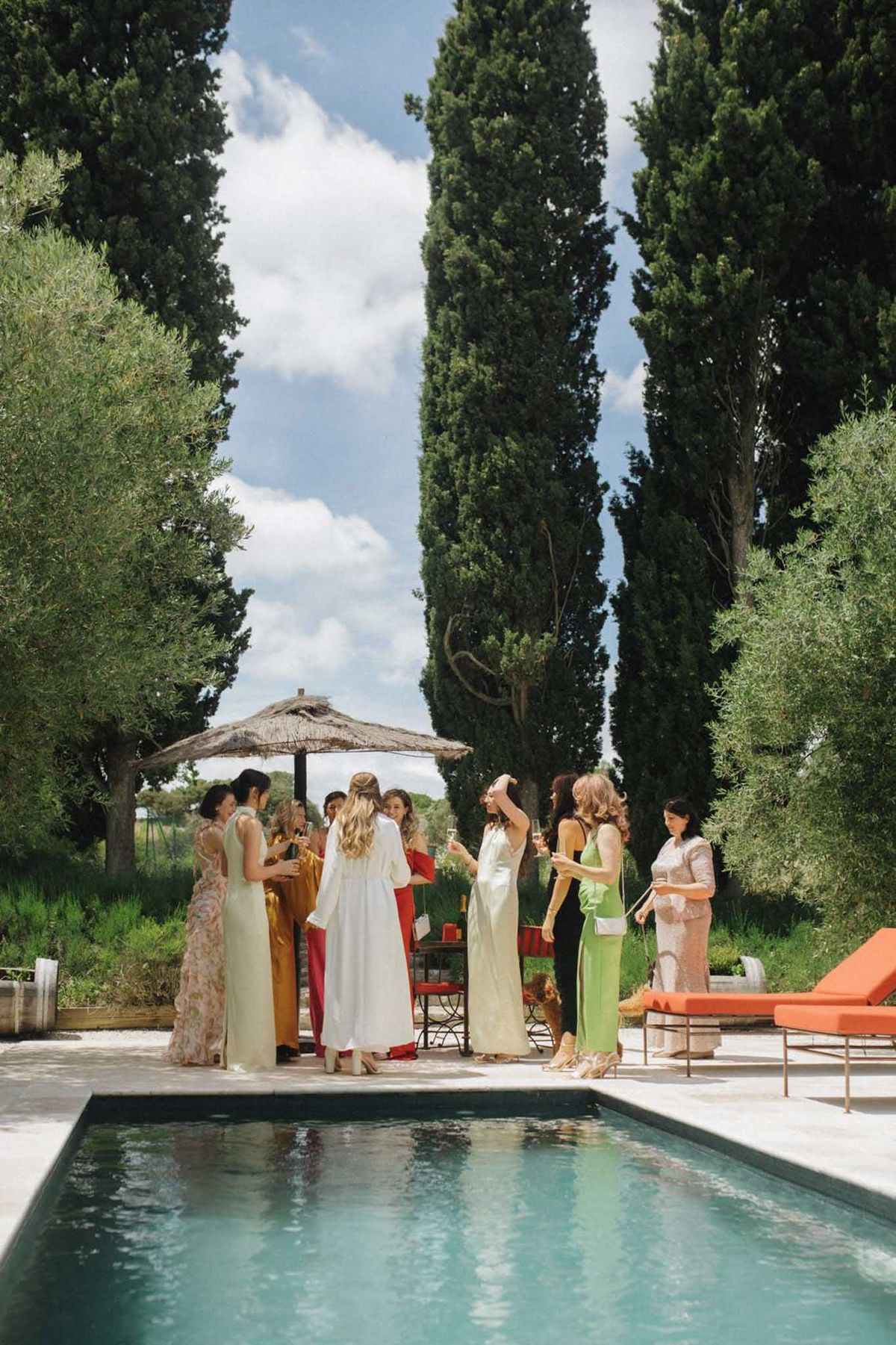 A getting-ready or pre-wedding gathering scene taking place outdoors beside a rectangular pool, likely at a Provençal or Mediterranean property. A group of approximately eight to ten women stand in a loose circle on the pool terrace, holding champagne glasses; the bride is identifiable in a long white robe with her hair half-up, surrounded by bridesmaids and guests dressed in a mix of floor-length gowns and separates in pale yellow, sage green, mustard gold, red, and black. A natural wicker parasol shades a small seating area with wrought-iron chairs in the background, and orange-cushioned sun loungers are visible to the right. The styling has a relaxed, summery feel with mismatched guest outfits suggesting an intentional colorful palette rather than a coordinated bridesmaid look. The shot is a medium-wide portrait taken from poolside level, with the group positioned at mid-distance.