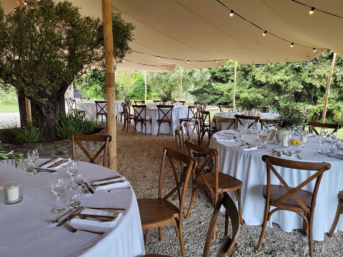 A wedding reception setup photographed from a medium-wide angle beneath an open-sided cream canvas stretch tent in a garden setting. Multiple round tables are dressed with white floor-length tablecloths and set with silver cutlery, white folded napkins, and clear wine glasses; the nearest table features a small white candle in a glass holder and a sprig of olive leaves as a simple centrepiece, while a table to the right displays yellow lemons and greenery as decoration. Seating is provided by dark walnut-stained cross-back wooden chairs arranged around each table, and the floor is loose gravel. Edison-bulb string lights are strung across the interior of the tent, adding warm overhead lighting. The overall styling is rustic-Provençal with a natural, understated palette of white linens, warm wood tones, and greenery. No guests are present, indicating the image was taken before the reception began.