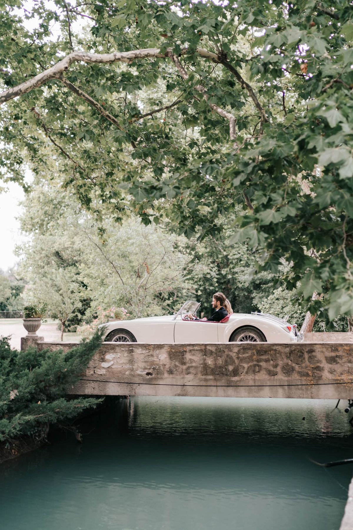 A man, likely the groom, is driving a cream-colored vintage open-top sports car — possibly a Jaguar XK — across a stone bridge over a calm teal-green waterway. He is dressed in a dark jacket and the car features a burgundy interior. The wide shot is taken from a slight distance, framed by overhanging tree branches, with a stone balustrade bridge in the foreground and lush estate grounds visible in the background. A decorative stone urn planter is visible on the bridge parapet, suggesting a classic French château or manor property setting.