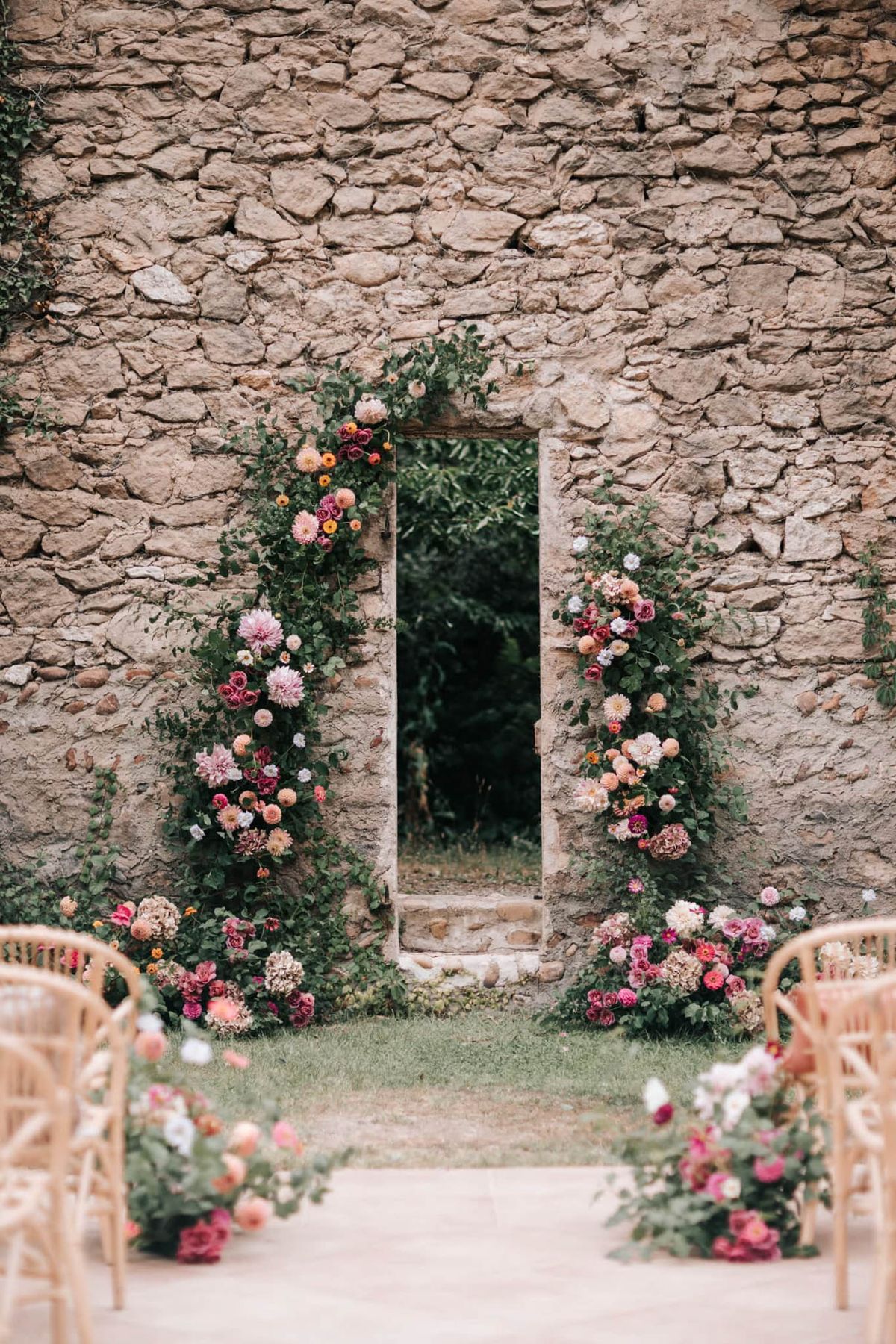 An outdoor wedding ceremony setup photographed from the aisle looking toward the altar, with no people present. The focal point is a tall, narrow stone doorway set into a rustic dry-stone wall, framed on both sides by lush floral installations featuring dahlias in shades of coral, blush, hot pink, burgundy, and peach, interspersed with roses, ranunculus, and trailing greenery including eucalyptus and climbing vines. Additional flower clusters are arranged at ground level on either side of the doorway and along the base of the aisle. Natural rattan or cane chairs are partially visible in soft focus on both sides of the aisle in the foreground. The overall decor palette is warm and richly colorful with a garden-inspired, romantic rustic aesthetic. Wide shot composition centered on the doorway arch. Potential venue feature image.