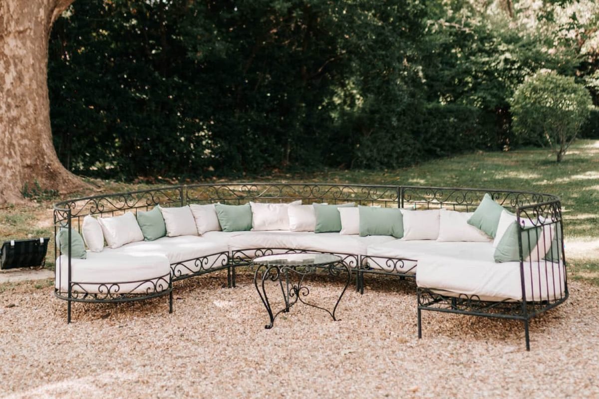 An outdoor lounge seating area set up for a wedding cocktail hour or reception, photographed as a wide decor detail shot. The seating consists of a large curved sectional sofa with a black wrought-iron scrollwork frame, fitted with cream/off-white cushions and accented with sage green throw pillows. A matching wrought-iron coffee table with a glass top sits at the center of the curved arrangement. The setup is positioned on a gravel surface in a garden setting, with no people present. The decor palette of cream and sage green reflects a classic French garden styling aesthetic.