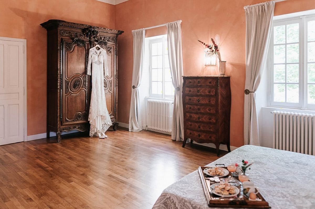 Lace bridal gown hangs on carved armoire with breakfast tray on bed in terracotta-walled chateau room