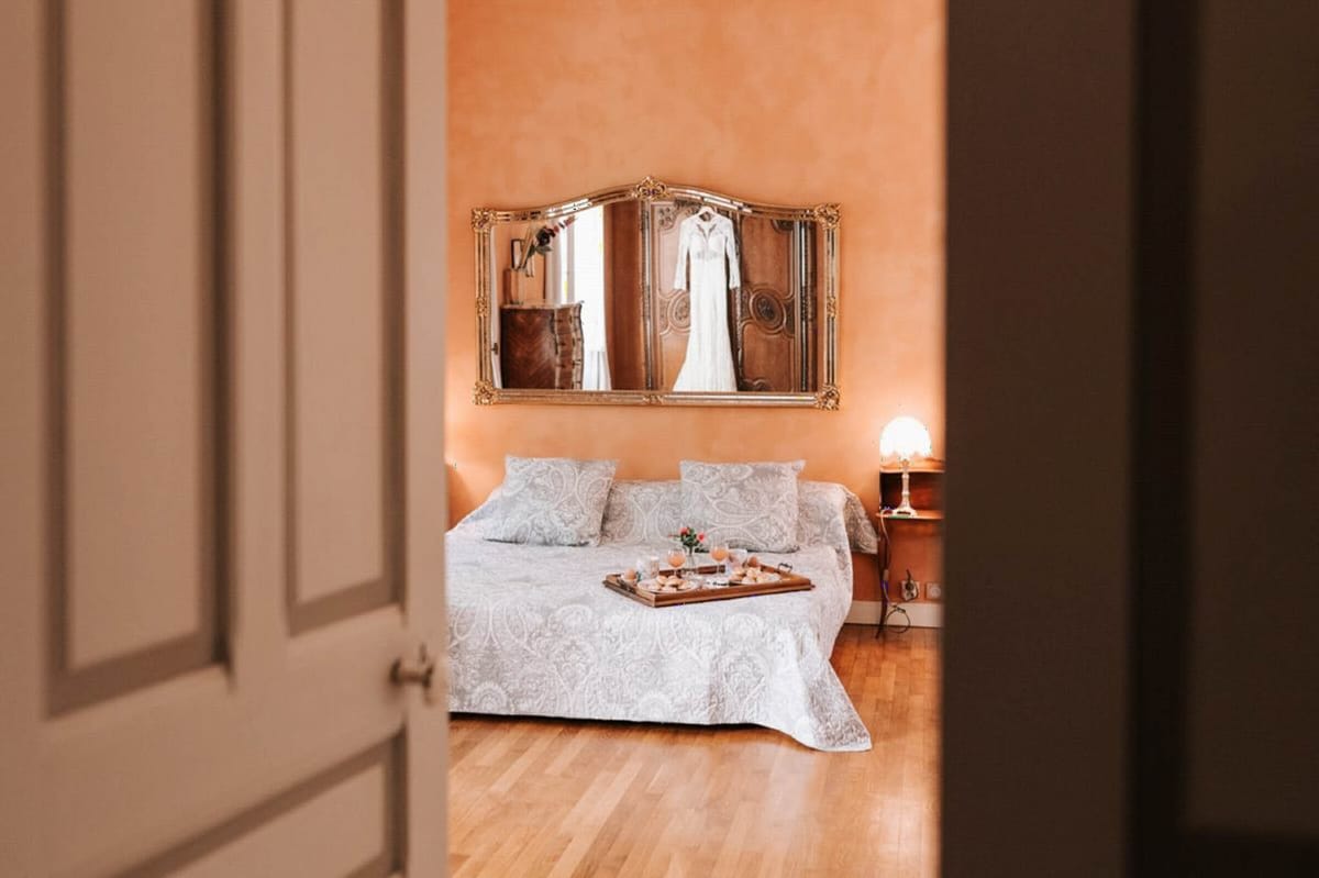 A getting-ready scene shot from a doorway, looking into a warmly lit bedroom with terracotta-orange walls and parquet flooring. A white, long-sleeved wedding dress is visible hanging in the reflection of a gold ornate triptych mirror mounted above the bed. The bed is dressed in a white and grey paisley coverlet with matching pillows, and a wooden tray sits on top holding what appear to be drinks with orange-tinted glasses, small pastries or macarons, and a small pink flower. A bedside lamp provides soft warm light to the right. The composition is framed by the open white panelled door in the foreground, creating a peek-through perspective into the room.