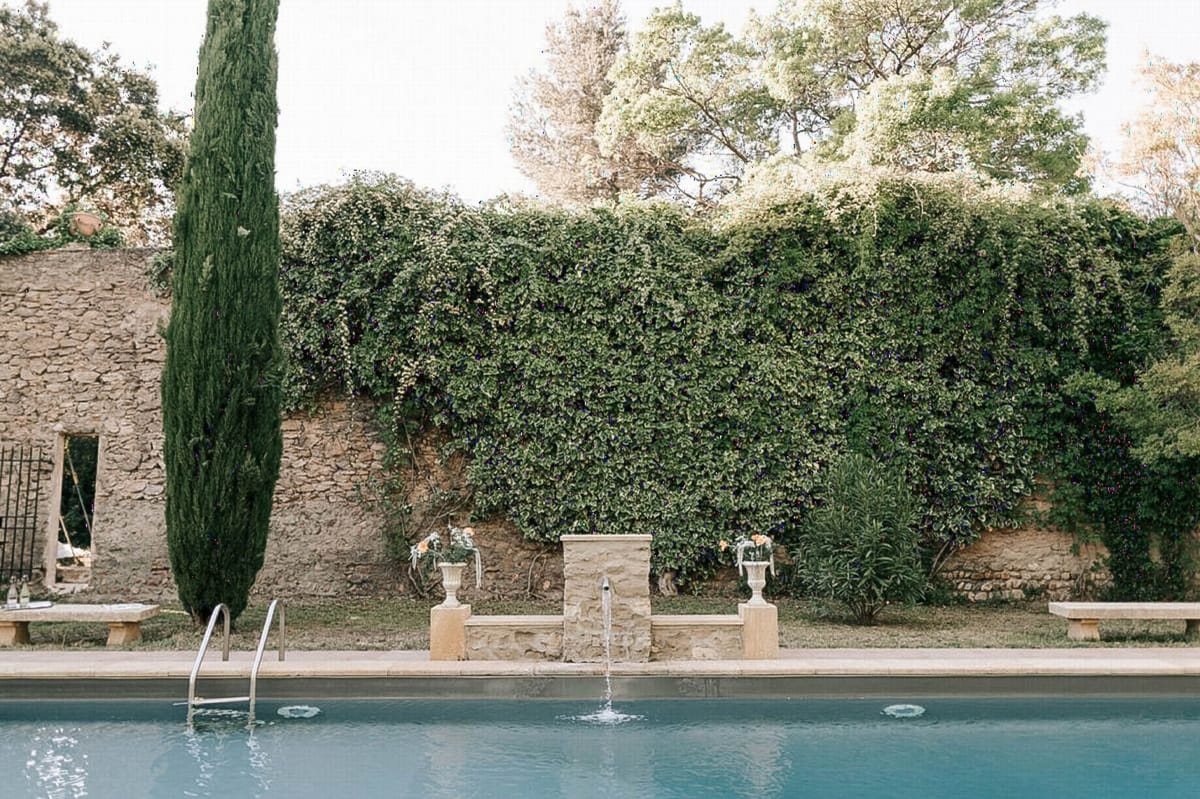 A wide shot of an outdoor pool area at what appears to be a French country estate or mas, with no people visible. The pool features a stone fountain spout at its center, flanked by two classical stone urns holding small yellow flower arrangements, set on stone pedestals. Two long stone benches run along the poolside, and a metal pool ladder is visible on the left. A dense ivy-covered wall forms the backdrop, with a tall cypress tree and an old ruined stone structure visible to the left. The styling is classic Provençal, with the decorative urns and yellow blooms adding a deliberate wedding touch to the venue grounds. Potential venue feature image.