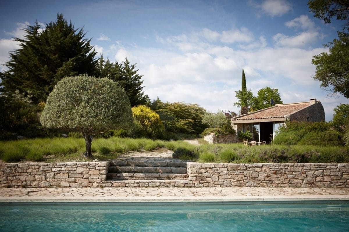 Rectangular pool with stone retaining wall and staircase before Provencal stone building with terracotta roof