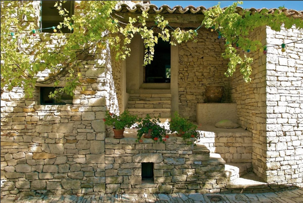 Outdoor wide shot of a traditional Provençal stone building exterior featuring a covered entrance with stone steps leading inward, terracotta roof tiles, and dry-stacked limestone walls. Terra cotta pots with red geraniums are arranged along a low stone ledge at the base of the entrance steps. Colorful globe string lights in yellow, red, and green are strung horizontally across the facade, suggesting event decoration. A climbing vine with green leaves trails across the upper portion of the entrance archway. No people are visible in the frame. Potential venue feature image.
