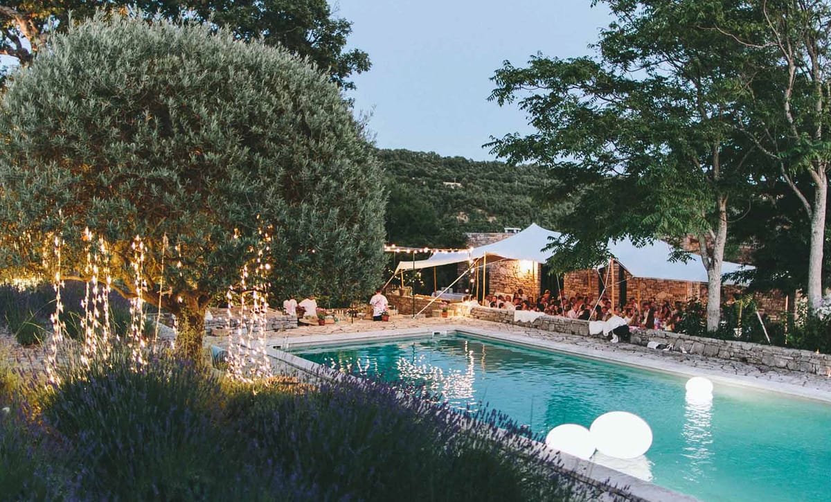 An outdoor evening reception scene photographed from a wide angle, showing a pool terrace at dusk at what appears to be a French stone-walled property. A large group of guests — approximately 30 to 50 people — are seated under two white stretch tent canopies positioned against a rustic stone wall. String lights are strung between the tents and surrounding trees, adding warm golden lighting to the scene. Cascading fairy lights hang from an olive tree on the left side of the pool, and two glowing white spherical float lights sit on the surface of the turquoise pool in the foreground. Lavender plants line the near edge of the pool terrace. The overall decor palette is white and warm amber, with a relaxed, natural style. Potential venue feature image.