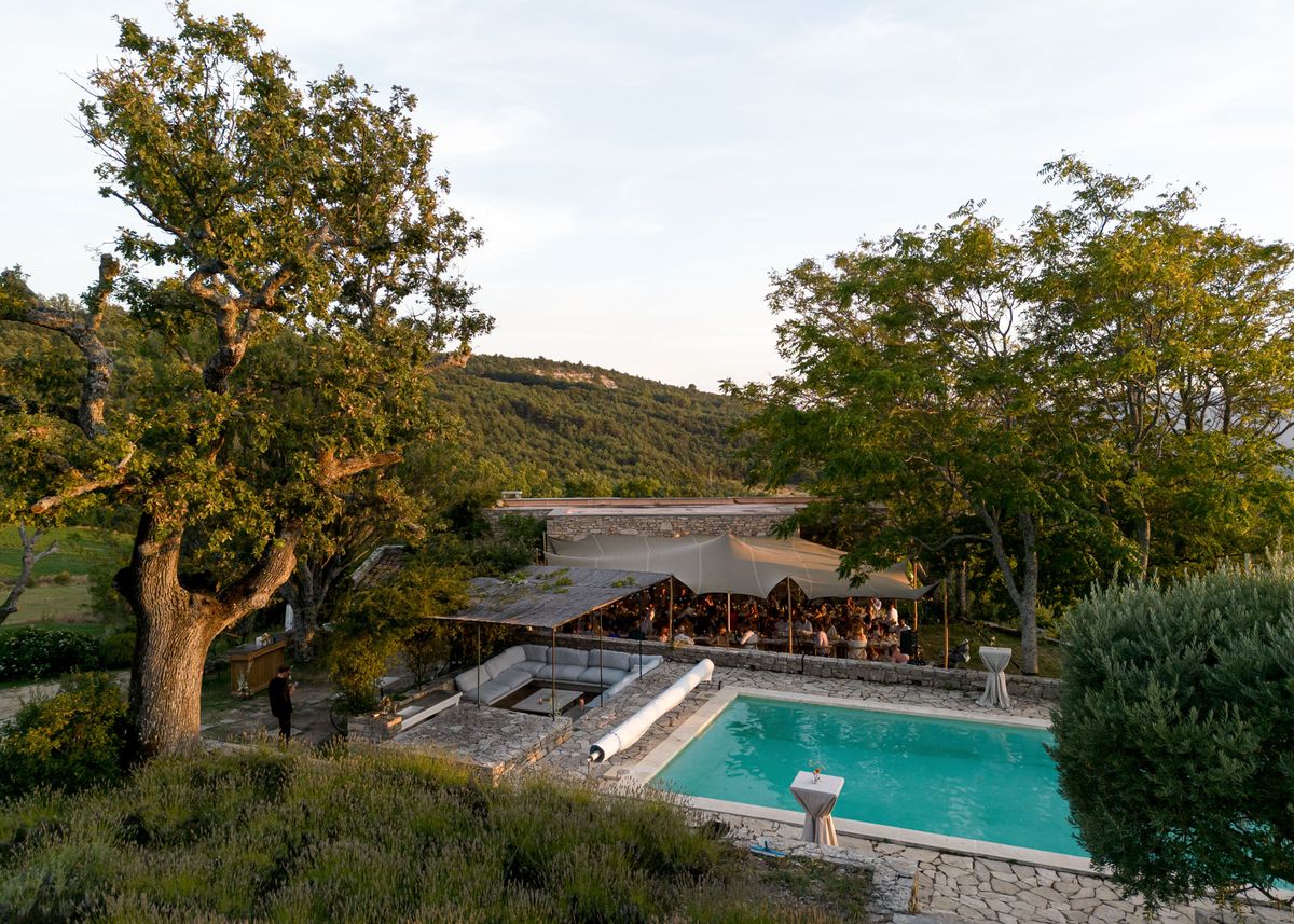 An aerial wide shot captures an outdoor wedding reception in progress at a stone-built property in a hilly, wooded landscape typical of the south of France. A large stretch tent with a warm golden-beige canopy covers a seated reception dinner for what appears to be 80–100 guests, with visible warm candlelight and festoon lighting glowing beneath it. Adjacent to the reception area is a stone terrace featuring a rectangular swimming pool with turquoise water, flanked by cocktail tables dressed in white linens, and a modern outdoor lounge area with grey sectional seating. The overall decor style is modern and understated, with a neutral palette of cream, grey, and natural stone tones. Potential venue feature image.