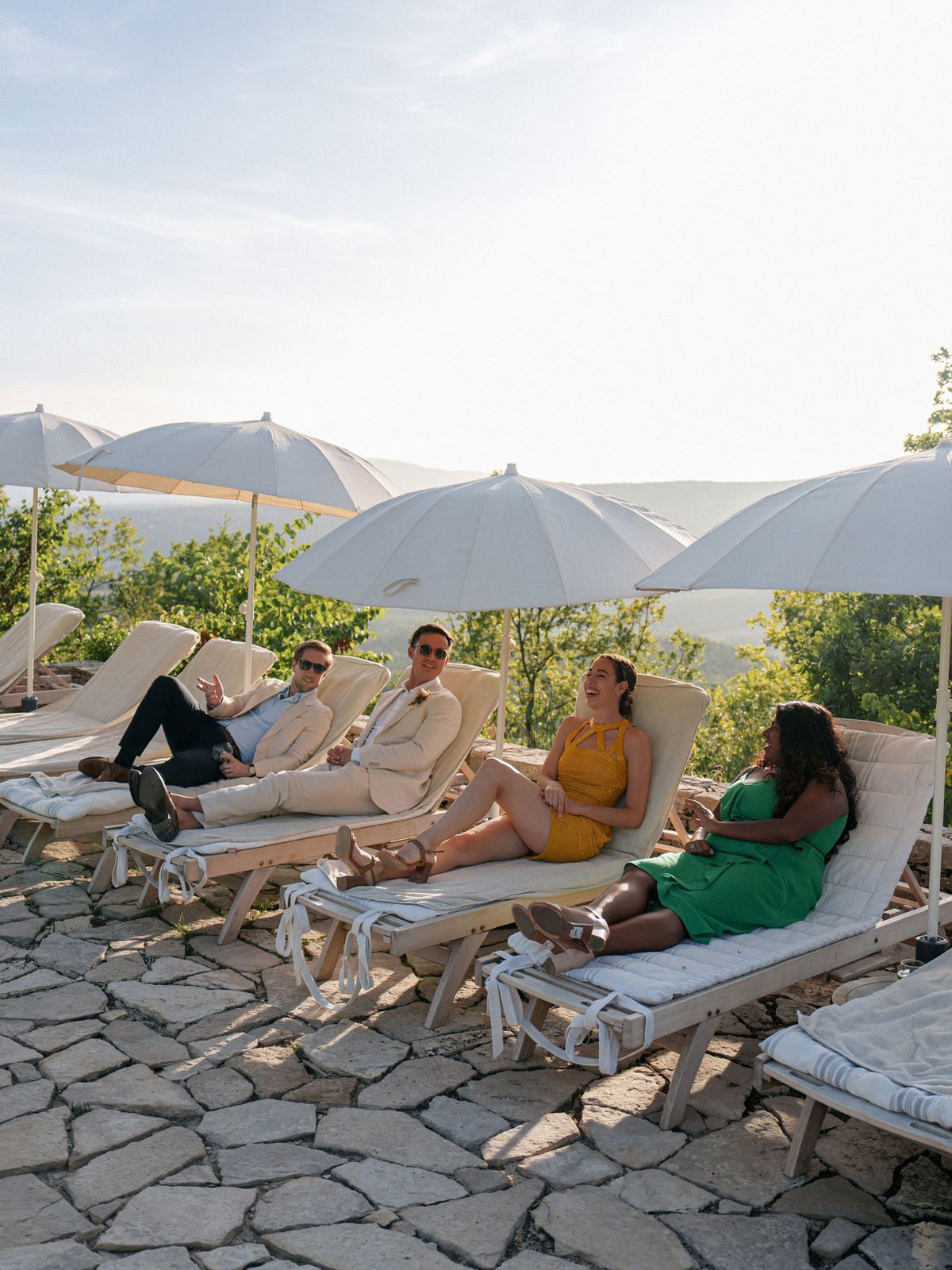 Four wedding guests relax on natural wood sun loungers with cream cushions during what appears to be a cocktail hour or outdoor reception break. The setting is an outdoor stone-paved terrace with a panoramic hillside view, shaded by several large white canvas market umbrellas. Two men are dressed in light linen suits — one sand-beige, one light blue — and two women wear a mustard yellow halterneck mini dress and a bright kelly green dress respectively. The group is laughing and conversing in a relaxed atmosphere. The wide shot captures the full terrace setup and the rolling wooded landscape in the background, suggesting an elevated countryside or domaine venue in southern France.
