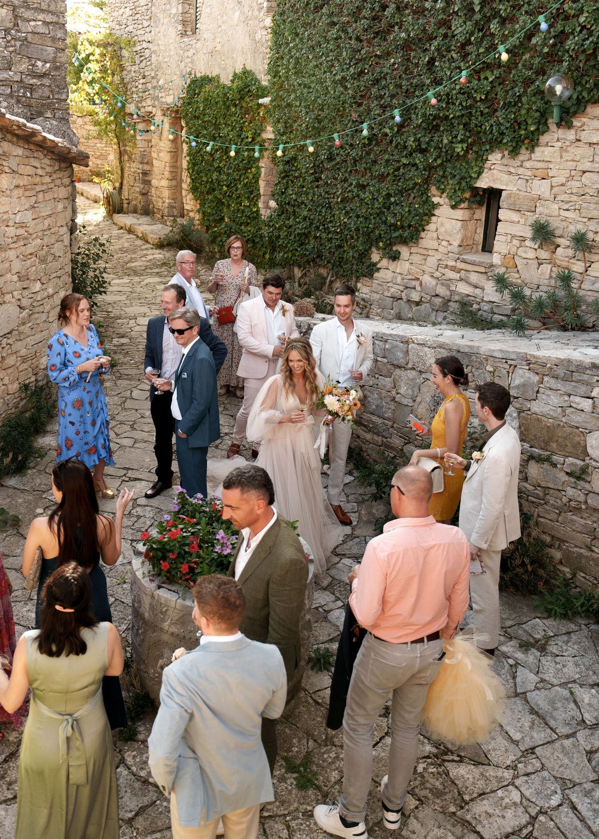 A cocktail hour scene in a narrow cobblestone courtyard or alleyway flanked by old stone walls, with ivy climbing one wall and a string of colorful festoon bulb lights strung overhead. Approximately 15 guests and the couple are mingling, holding drinks, in a relaxed outdoor setting. The bride wears a floaty, sheer blush-toned gown with long tulle sleeves and a flower crown, holding a loose bouquet of warm-toned blooms including white dahlias and orange and peach flowers. Guests are dressed in a mix of summer attire — a mustard yellow dress, sage green dress, olive suit, blush pink blazer, and navy and blue patterned outfits — giving the gathering a relaxed, boho-rustic feel. The shot is taken from an elevated angle, providing a wide overhead view of the full scene.