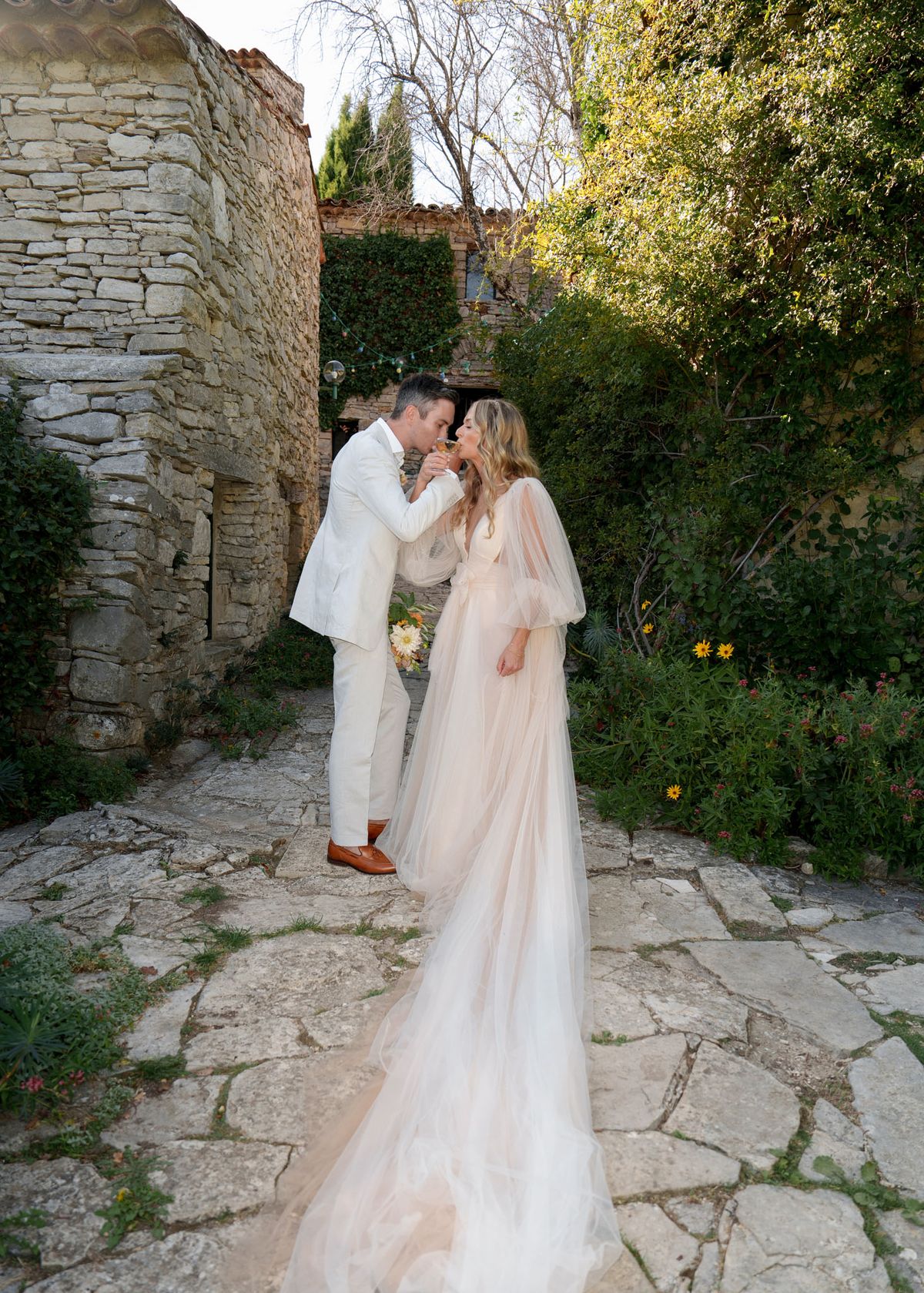 Couple sharing arm-linked champagne toast in stone courtyard, bride in blush tulle gown with cape sleeves