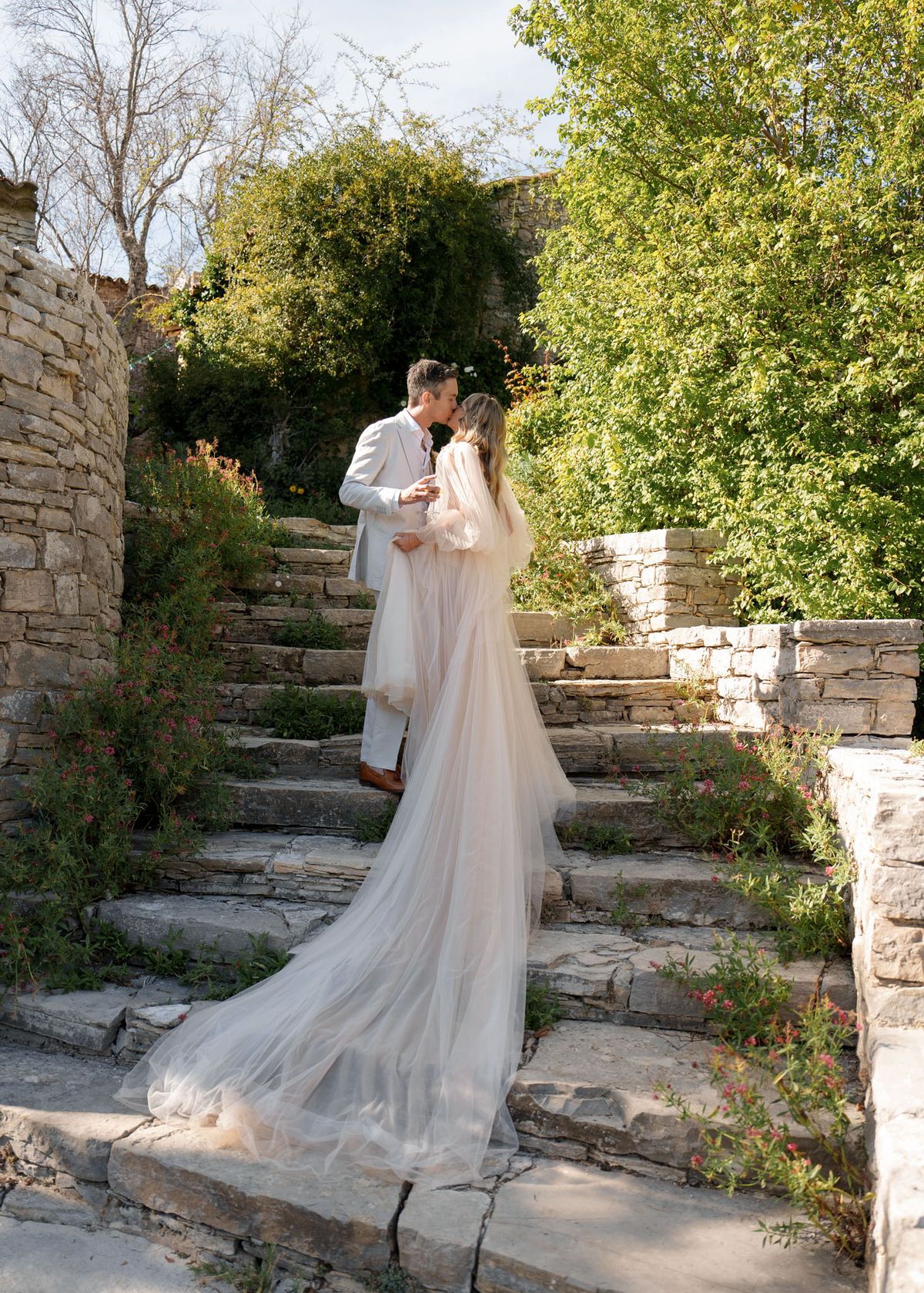 A couple shares a kiss during an outdoor portrait session on a wide stone staircase flanked by rustic dry-stone walls. The bride wears a long-sleeved ivory tulle gown with a voluminous cathedral-length train that cascades down several steps, styled in a romantic boho aesthetic. The groom wears a light grey suit with brown shoes. The setting appears to be a terraced garden with mature trees and scattered wildflowers in pink and orange tones growing between the stonework. The shot is taken from behind and slightly below the couple, giving full emphasis to the dress train and the stone architecture surrounding them.