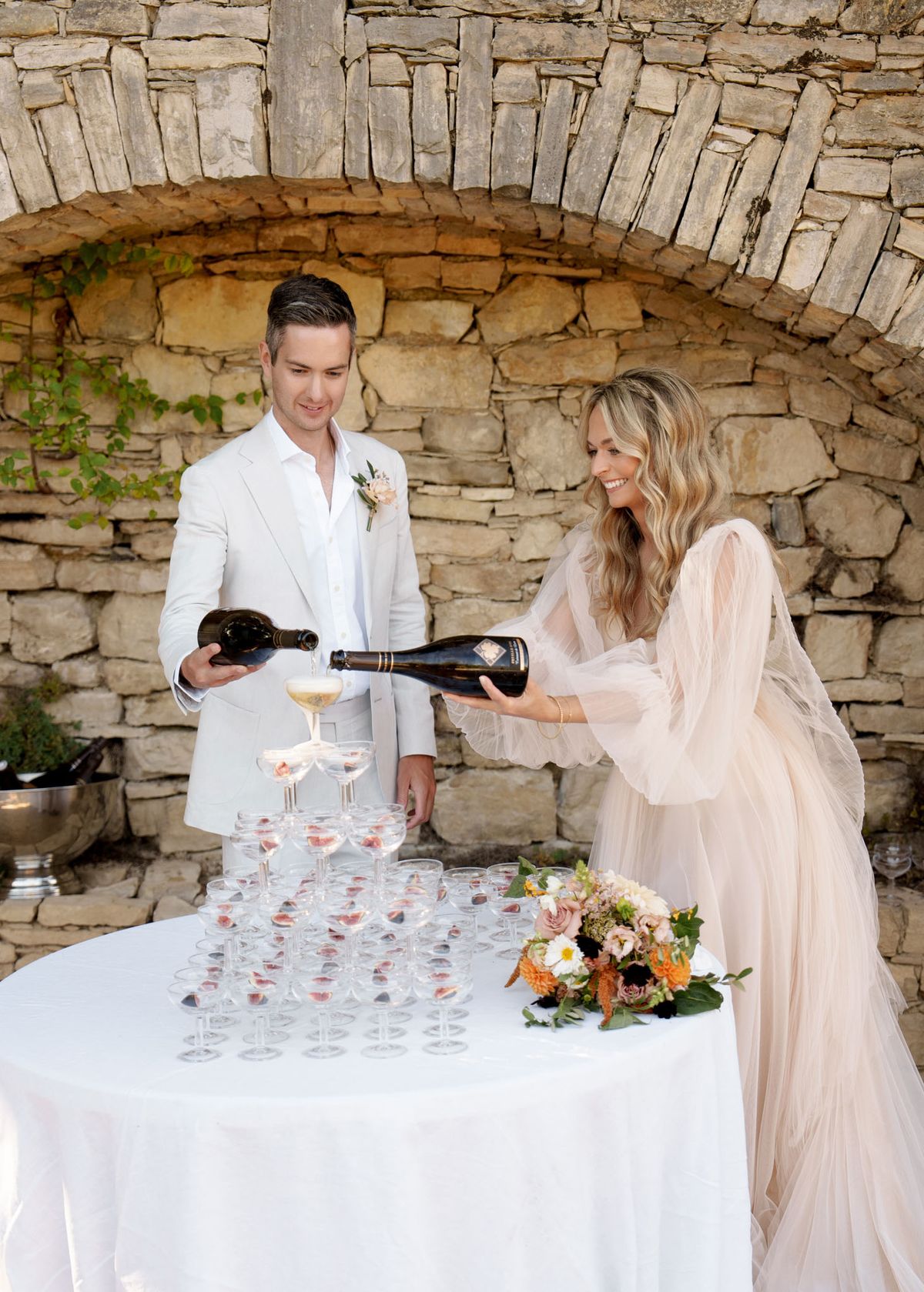 The bride and groom are pouring champagne together into a tiered tower of coupe glasses arranged on a white-linen-covered table, set against a rustic dry-stone arch backdrop outdoors. The groom wears an ivory suit with an open-collar white shirt and a blush boutonnière, while the bride wears a blush-toned gown with dramatic sheer, ruffled sleeves and flowing tulle overlay. Each coupe glass contains what appears to be a small piece of fruit, likely strawberries or figs. A mixed bouquet of orange dahlias, blush roses, white anemones, and greenery rests on the table beside the champagne tower, and a silver champagne bucket is visible to the left. The shot is a medium portrait-style image capturing both figures and the full table setup, with a boho-romantic styling direction.
