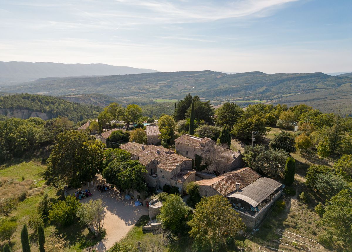 An aerial drone shot captures a large Provençal stone farmhouse complex with terracotta-tiled roofs set among dense trees, with a wide valley and mountain range visible in the background. The property comprises several interconnected stone buildings arranged around a gravel courtyard, with a covered barn structure visible to the right and a swimming pool visible further back on the grounds. A group of approximately 30–40 wedding guests can be seen gathered in the courtyard to the left of the main building, suggesting a ceremony or cocktail hour in progress outdoors. The wide establishing shot showcases the full scale of the venue, its rural Provençal architecture, and its elevated hillside position. Potential venue feature image.