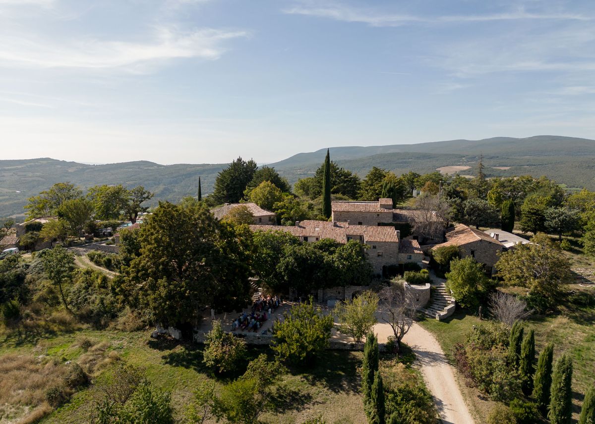 An aerial drone shot captures a wedding ceremony or gathering of approximately 20–30 guests assembled on an outdoor terrace or courtyard in front of a large stone mas-style property with terracotta-tiled roofs, typical of Provençal architecture. The venue is a multi-building stone estate set on an elevated hillside, surrounded by cypress trees, mature oaks, and manicured grounds, with a gravel driveway visible to the right. The guests appear to be seated or standing in a loose group on the lower terrace area, suggesting an outdoor ceremony or cocktail moment in progress. The wide-angle aerial composition emphasizes the full scale of the property and its hillside setting, with rolling mountain ridges visible in the background. Potential venue feature image.
