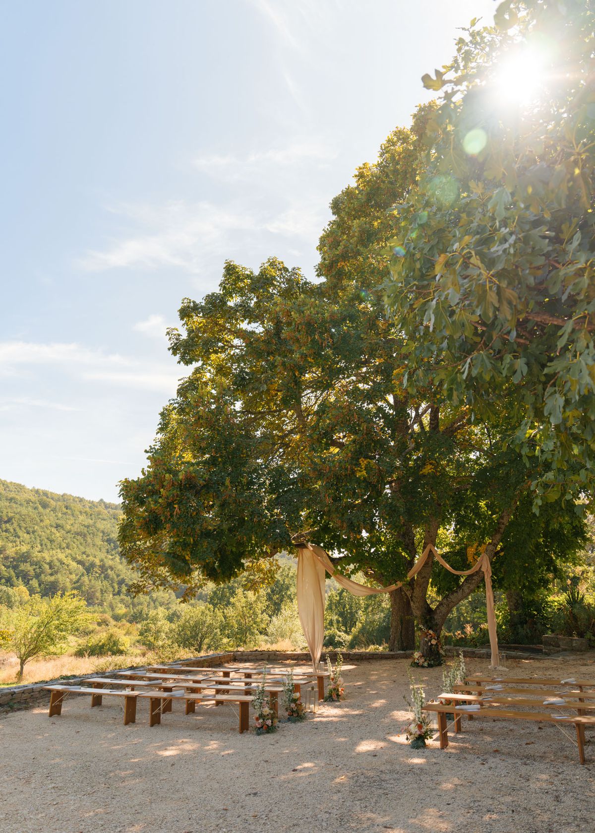 An outdoor ceremony setup photographed before the wedding, set on a gravel terrace in a rural landscape with wooded hillsides in the background. The ceremony space features rows of wooden benches arranged in two sections with a central aisle, framed by a draped arch made of warm ivory or champagne-toned fabric tied around the trunk and branches of a large mature tree. Small floral arrangements in coral, peach, and white tones are placed at the ends of select benches and at the base of the arch. A tall glass lantern is visible near the altar area. The styling is rustic and natural, using the tree itself as the central focal point of the altar. Wide shot taken from behind the seating area, with strong sun flare from the upper right corner. Potential venue feature image.