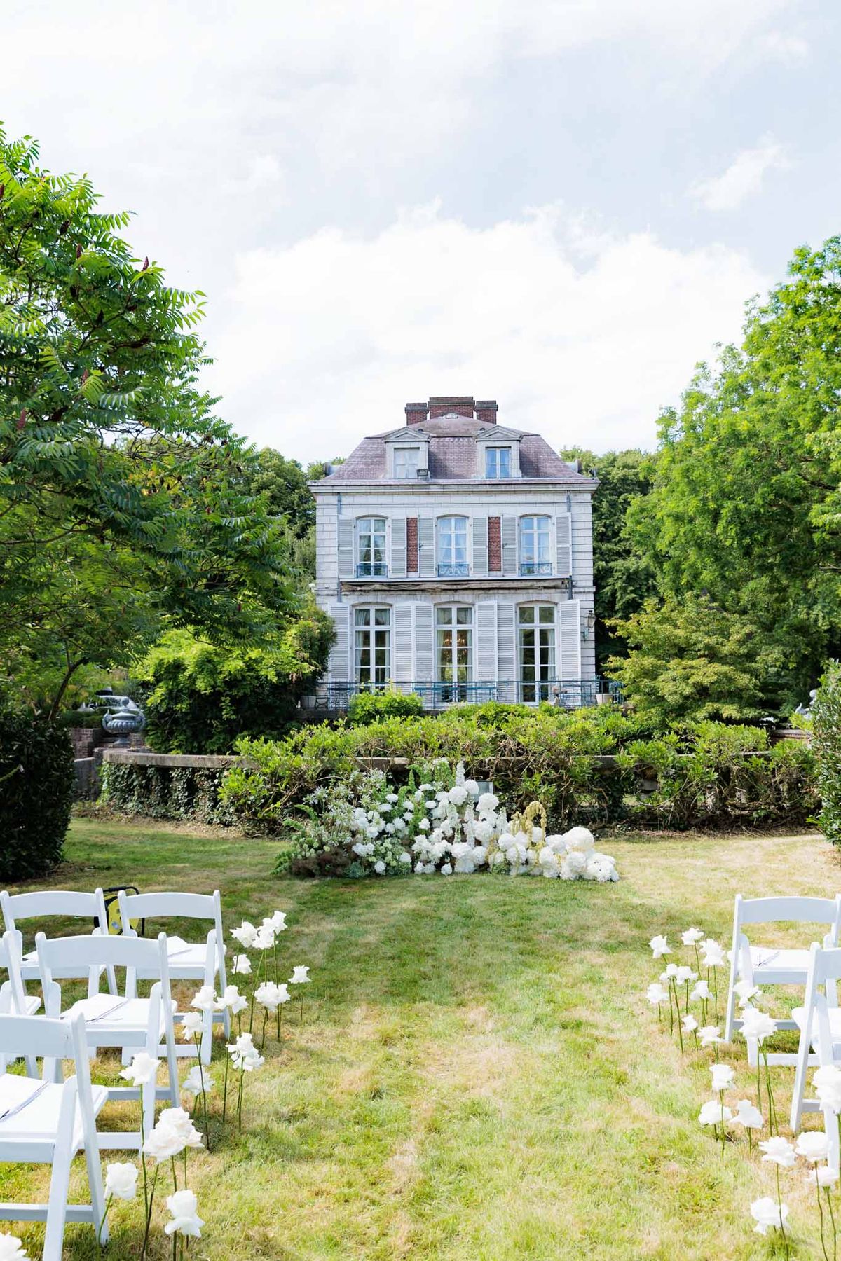 An outdoor wedding ceremony setup on the lawn of a French manor house, photographed before guests have arrived. White folding chairs are arranged in rows on either side of a grass aisle, with tall white florals on slender stems lining the aisle. The altar area features a large low arrangement of white blooms — including what appear to be white hydrangeas and peonies — placed directly on the ground in front of a low stone garden wall and hedgerow. The venue is a three-storey French manor with pale stone façade, mansard roof, brick chimney stacks, and white shuttered windows. The overall decor palette is all-white with a classic, garden-style aesthetic. Wide shot taken from behind the guest seating looking toward the altar and manor building. Potential venue feature image.