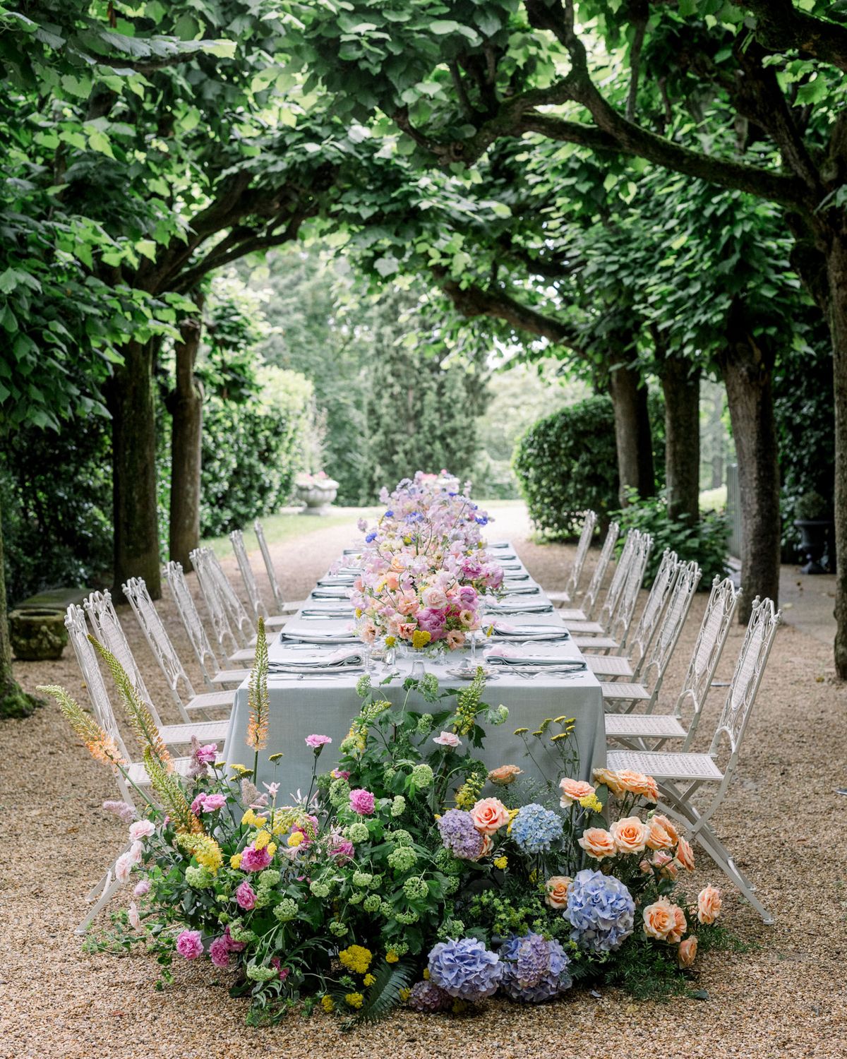 Outdoor reception table in garden allee with lush pink roses, blue hydrangeas, and purple delphiniums