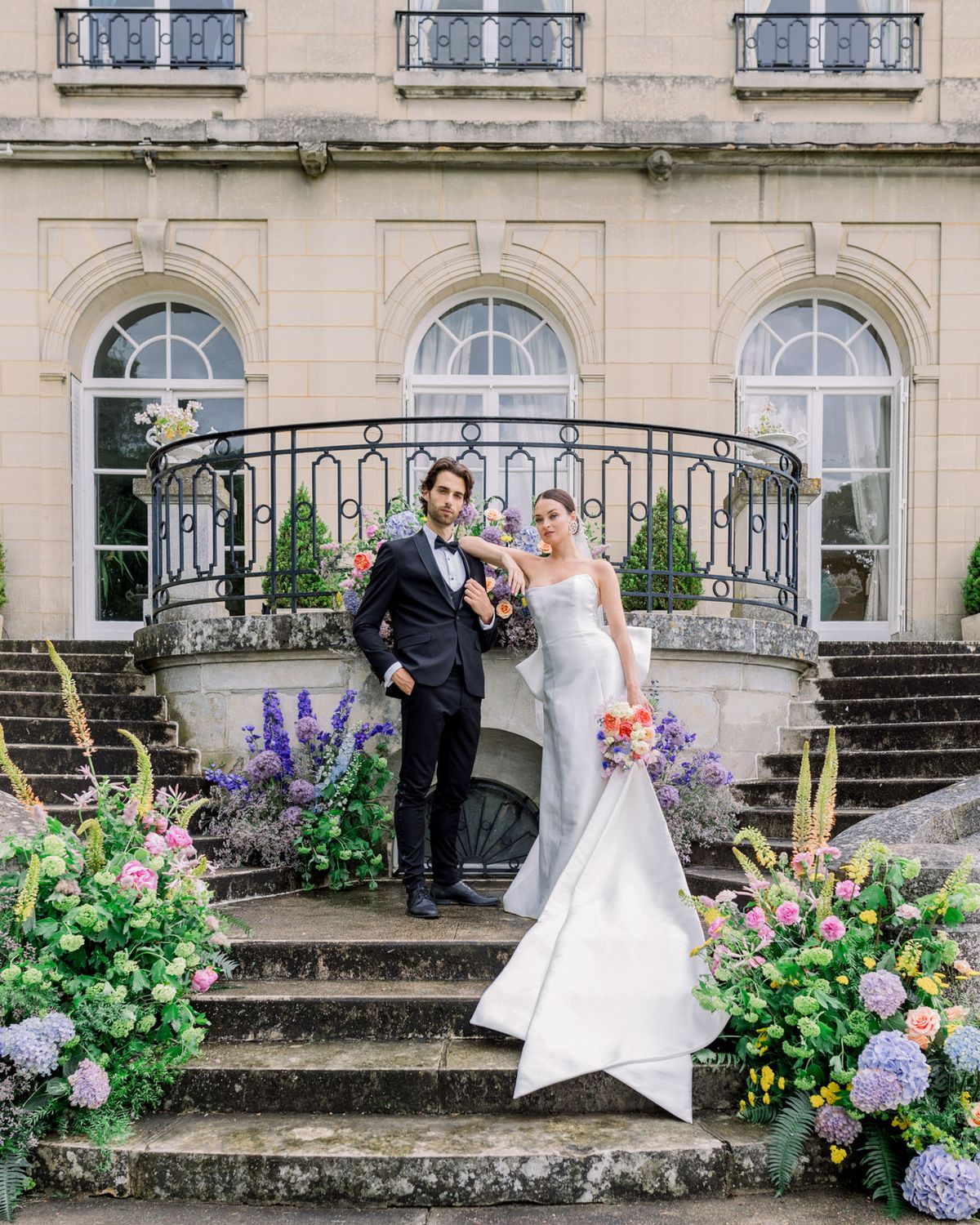 Bride and groom posing on grand stone staircase lined with coral roses and purple hydrangeas at French chateau
