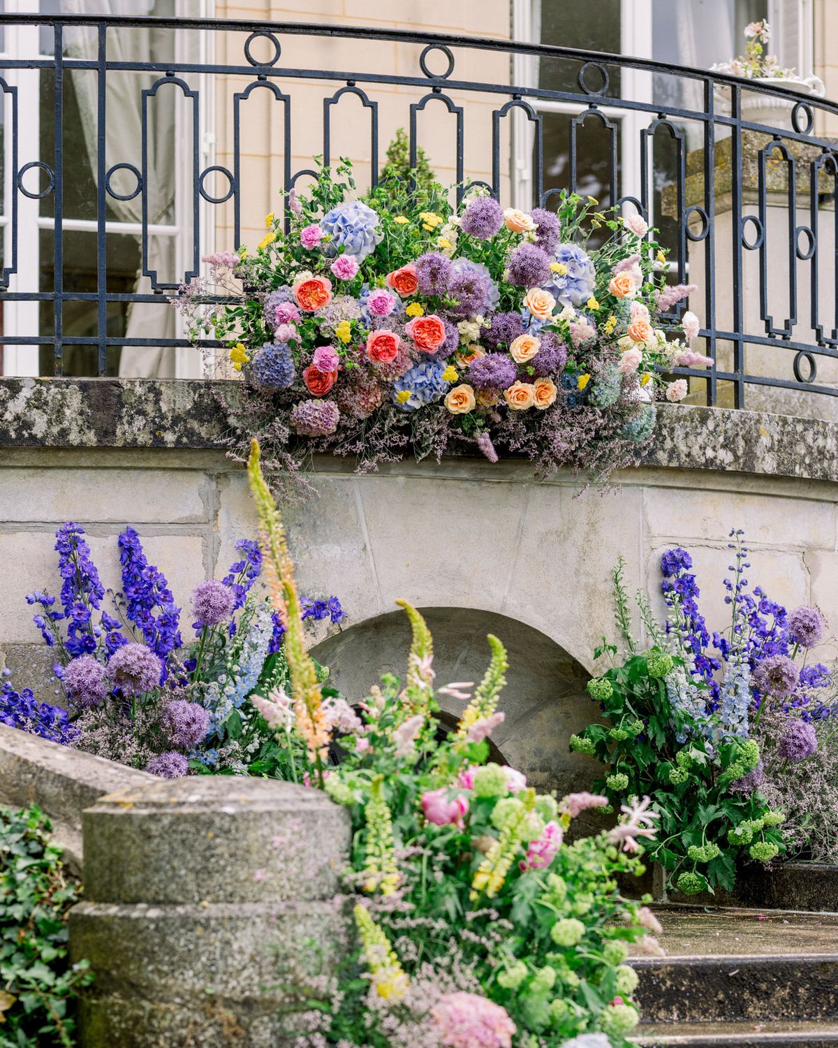Floral installation on classical stone archway with blue hydrangeas, coral roses, mauve dahlias, and purple delphiniums