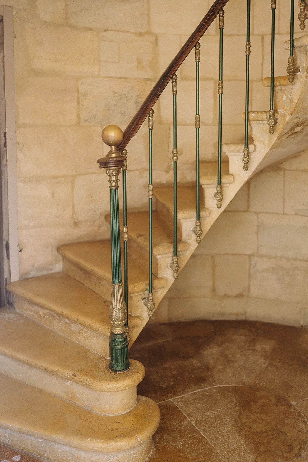 A detail shot of an interior stone staircase inside what appears to be a French château or historic manor. The staircase features carved limestone steps and a wrought iron balustrade painted in deep forest green with ornate gold-toned decorative fittings, including a spherical brass newel post cap and fluted balusters with carved detailing. The handrail is a warm dark wood. The surrounding walls are constructed from pale cream cut stone blocks, and the ground floor is laid with large irregular stone tiles in warm beige and brown tones. No people are present. The composition is a close-to-medium architectural detail shot taken from ground level. Potential venue feature image.