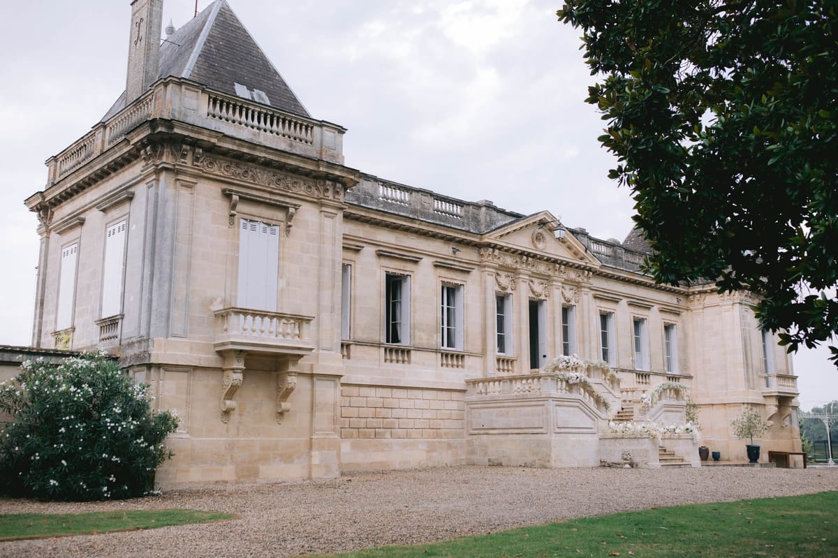 A wide exterior shot of a French château with classic 18th-century architecture, featuring cream limestone façades, ornate carved stonework, tall shuttered windows, a balustraded terrace, and a pointed slate-roofed tower on the left. The entrance staircase is decorated with arrangements of white flowers — appearing to be white roses and possibly baby's breath — draped along the balustrade and flanked by potted topiaries in dark planters, indicating the venue is dressed for a wedding. No people are visible in the frame. The gravel forecourt and manicured lawn are visible in the foreground. Potential venue feature image.
