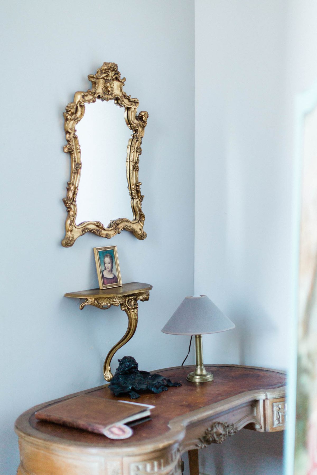 A detail interior shot of a room with pale blue-grey walls, featuring a gold rococo-style ornate mirror with cherub and scroll carvings hung above a matching small gold wall-mounted console shelf. On the shelf sits a small framed portrait painting of a woman in a classic style. Below, a curved antique walnut writing desk with carved floral detailing holds a brass table lamp with a conical grey shade and a small dark bronze animal figurine. A paper or card with a red wax seal is also visible on the desk surface. The composition is a close-to-medium interior shot with soft natural light entering from the right. Potential venue feature image.