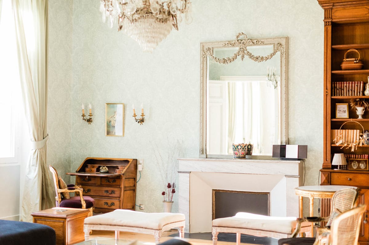 An interior room shot of what appears to be a French château or manor house, shown without any wedding activity in progress. The room features pale mint-green damask wallpaper, a crystal chandelier, and a white marble fireplace topped with an ornate carved white mirror with garland detailing. Furnishings include gilt-framed Louis XVI-style chairs with cream upholstered ottomans, a walnut roll-top secretary desk, gold brass wall sconces, and a tall wooden bookcase filled with leather-bound books and decorative objects. A small framed painting and a decorative vase with dried stems add additional detail to the space. The overall interior style is classic French period decor with a neutral ivory and warm wood palette. Wide interior shot with natural light entering from tall curtained windows at left. Potential venue feature image.