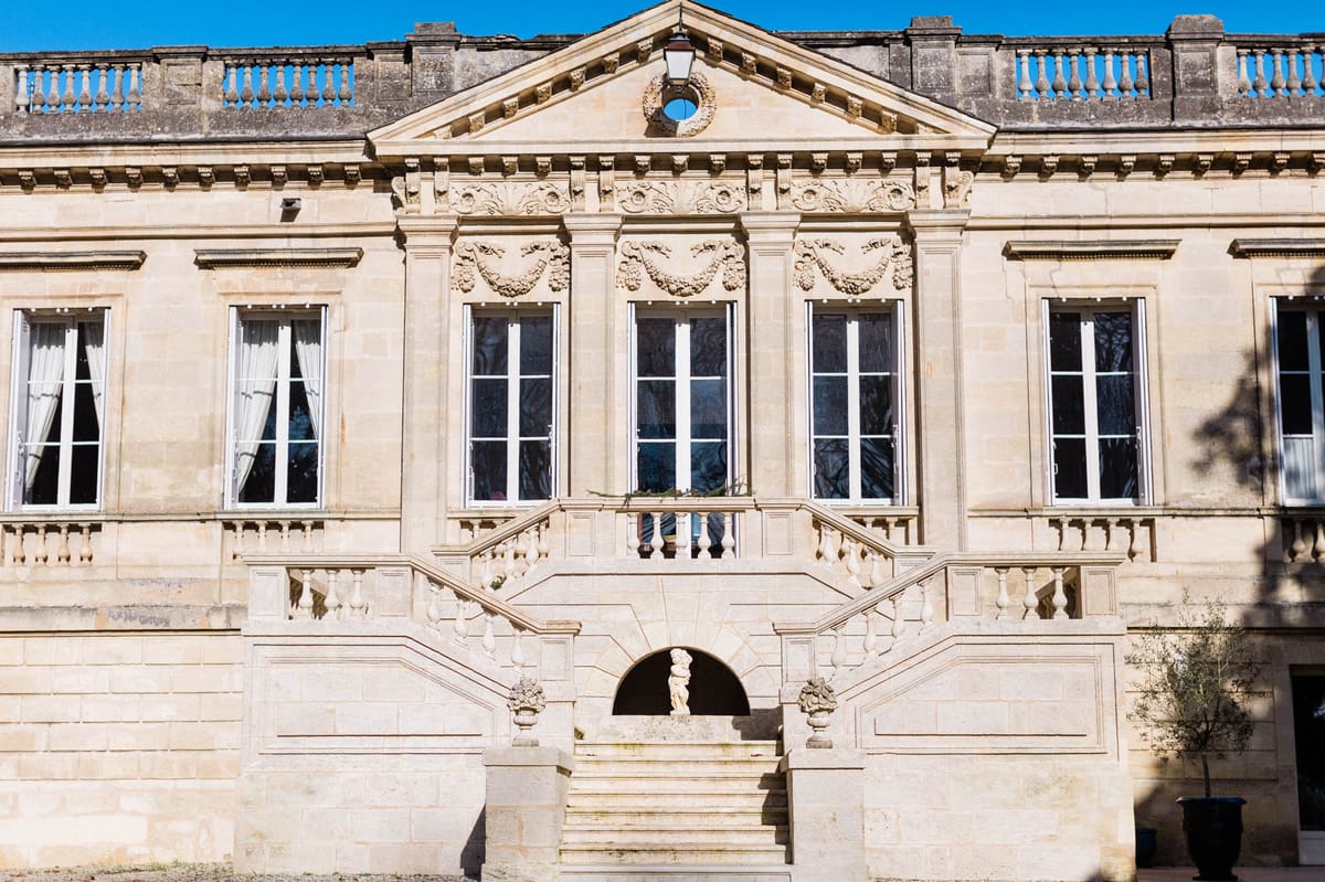 A wide exterior shot of a French classical château façade built in pale limestone. The building features a central pediment with decorative carved stonework including garland and wreath motifs, tall white-framed French windows across two visible floors, and a grand double staircase with balustraded railings leading up to a landing. A small classical stone statue is visible beneath an arched niche at the base of the staircase, and ornamental urns flank the steps. No people are visible in the image. The photograph is taken straight-on in bright daylight with no wedding-specific decor present. Potential venue feature image.