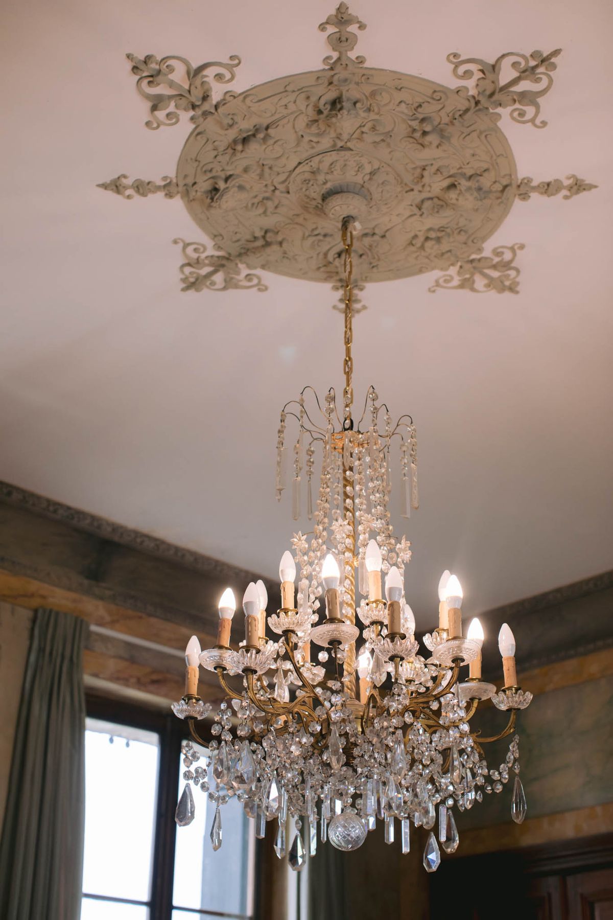 Detail shot looking up at a large multi-arm crystal and gilt brass chandelier with candle-style bulbs, suspended from an ornate plaster ceiling rose with scrollwork and acanthus leaf detailing painted in off-white. The chandelier features approximately 16 arms across two tiers, adorned with hanging crystal drops, prisms, and beaded strands. The interior space shows dark wood paneling or beams along the upper walls, grey-green curtains framing a tall window, and warm ambient light coming through the glass. The overall interior styling reflects a classic French château aesthetic. Potential venue feature image.