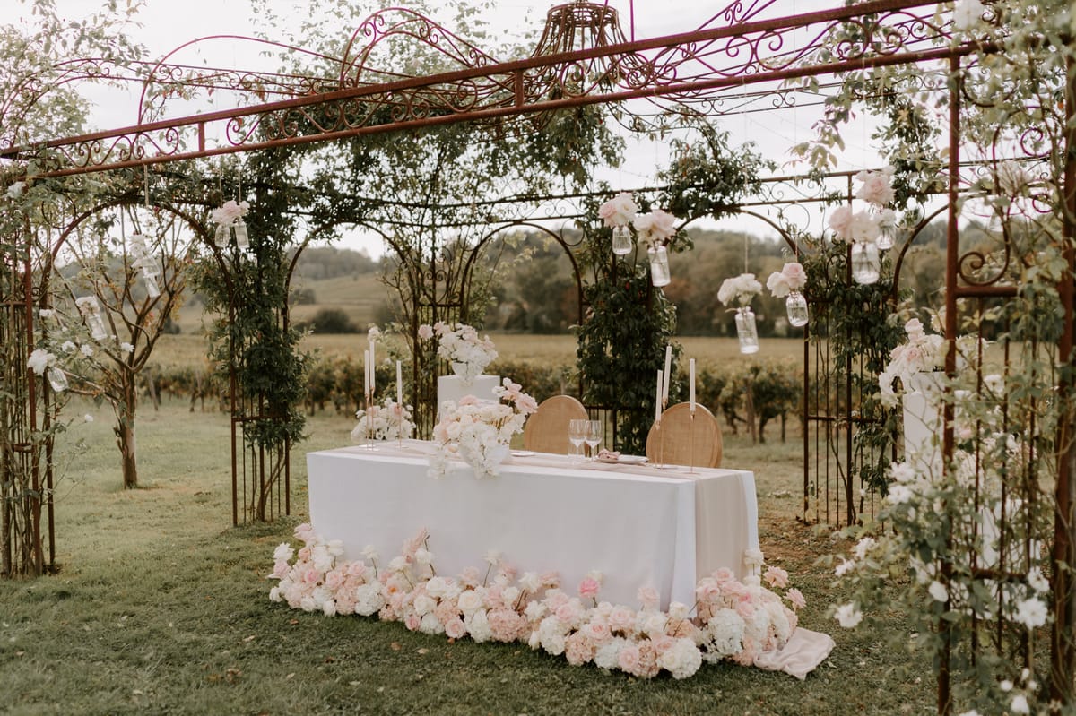Sweetheart table for two under iron pergola with blush peonies, taper candles, and vineyard backdrop