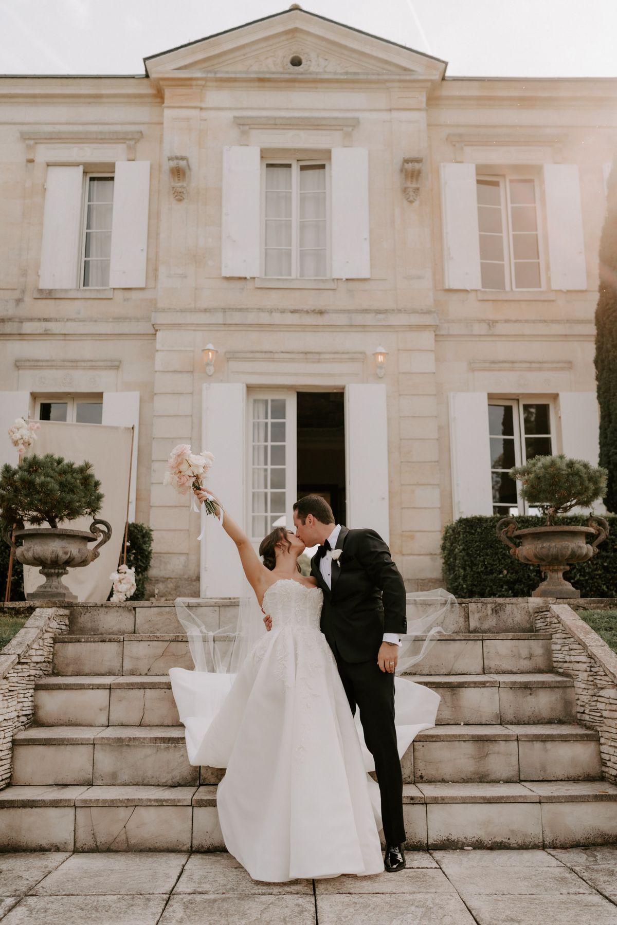 The bride and groom share a kiss on the stone entrance steps of a classic French château, captured in a full-length portrait shot. The bride wears a strapless ivory ballgown with a lace-detailed bodice and a long flowing veil that billows to the side, and she raises her bouquet of blush pink peonies and roses overhead with one arm extended. The groom is dressed in a black tuxedo with a white boutonnière and black bow tie. The château façade features pale limestone architecture with white shuttered windows, a triangular pediment, wall-mounted lanterns, and large ornamental stone urns with clipped topiary flanking the entrance steps. Potential venue feature image.