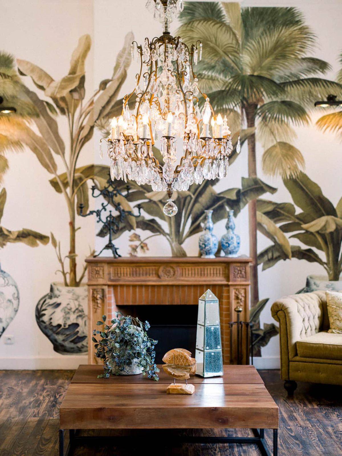 Interior lounge with tropical botanical wall mural, carved fireplace, brass chandelier, and tufted armchair