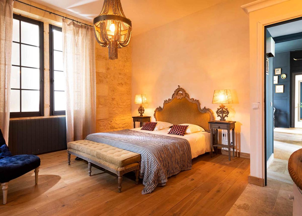 Interior shot of a bedroom, likely part of a château or manor house used as a wedding venue. The room features a double bed with an ornate carved wooden headboard upholstered in gold fabric, dressed with white bedding, a navy-and-white striped throw, and two red-and-black polka dot accent cushions. A tufted upholstered bench sits at the foot of the bed, and matching carved wooden bedside tables hold warm-toned table lamps on either side. The ceiling is fitted with a decorative beaded chandelier emitting warm amber light, and the floor is wide-plank oak hardwood. Exposed limestone stonework is visible on one wall alongside tall black-framed windows dressed with sheer linen curtains, and a navy velvet armchair is partially visible in the lower left corner. The adjoining room visible through the open doorway is painted deep navy blue. The overall decor style blends French antique and eclectic elements with warm, moody lighting. Potential venue feature image.