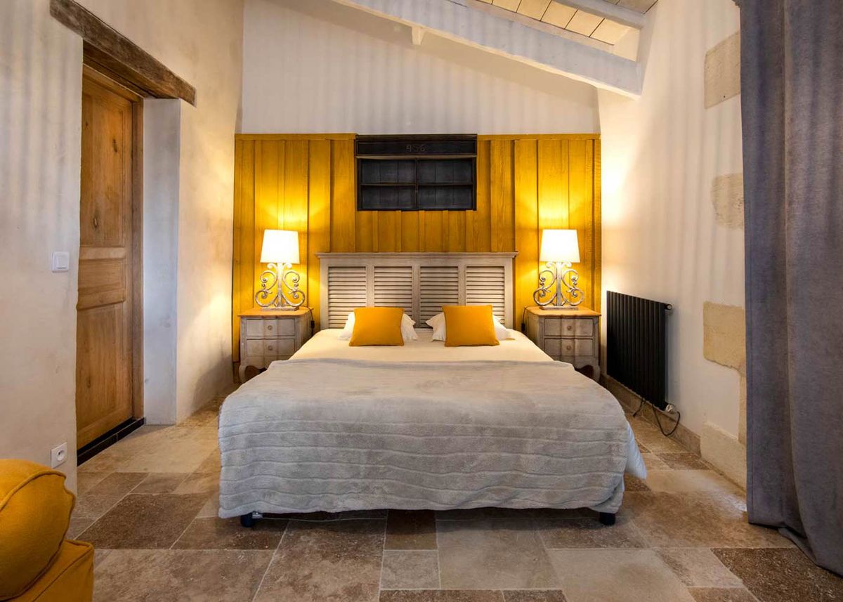 Interior shot of a guest bedroom at a French wedding venue, styled in a rustic-provincial aesthetic. The room features a double bed with a grey textured throw and white bedding, dressed with two mustard yellow cushions, set against a painted wood-panel headboard in a warm ochre-yellow tone. Two matching ornate wrought-iron bedside lamps with white shades flank the bed on distressed grey wooden nightstands, casting warm ambient light. The room has exposed ceiling beams, stone tile flooring, plastered walls with visible aged texture, a heavy grey linen curtain to the right, and a wooden door to the left — consistent with a renovated French farmhouse or mas interior. Potential venue feature image.