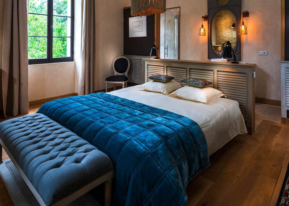 Guest bedroom in French chateau with teal velvet bedspread, grey tufted bench, and cage-style pendant lighting